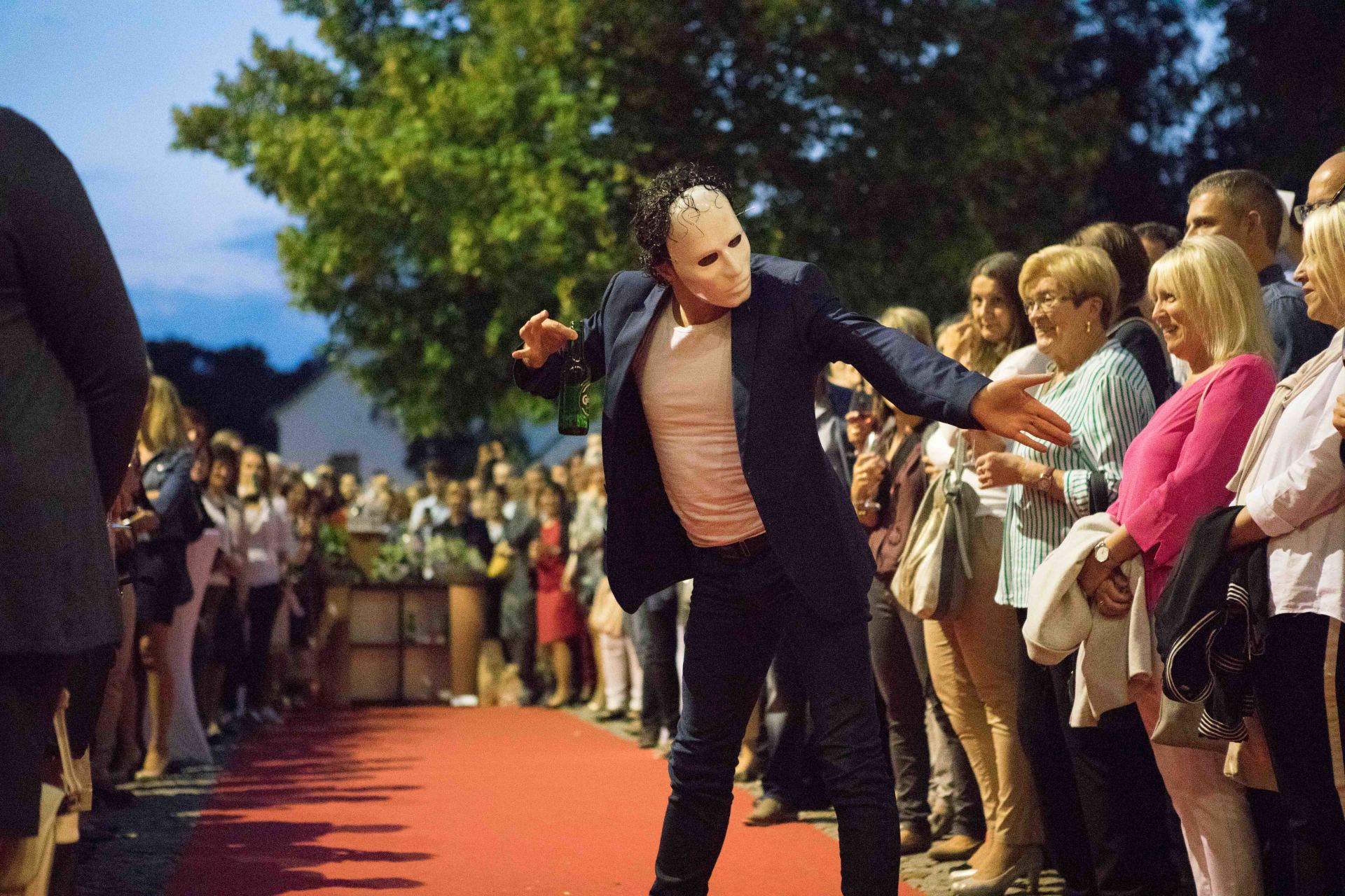 Ein Mann mit einer Maske tanzt vor einer Menschenmenge auf einem roten Teppich.