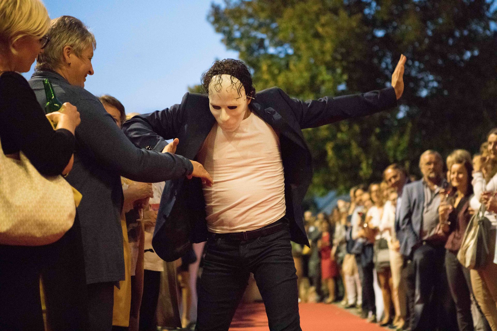 Ein Mann mit einer Maske tanzt vor einer Menschenmenge auf einem roten Teppich.
