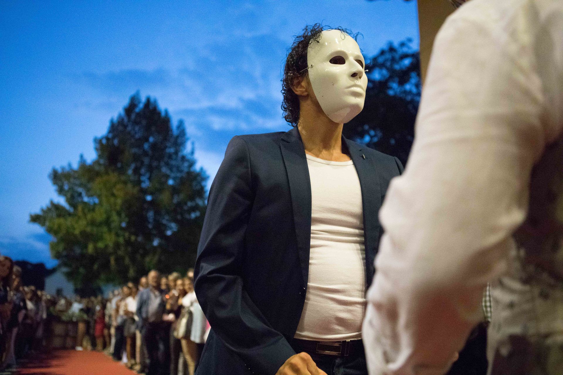 Ein Mann mit einer weißen Maske und einem Anzug steht neben einem anderen Mann.