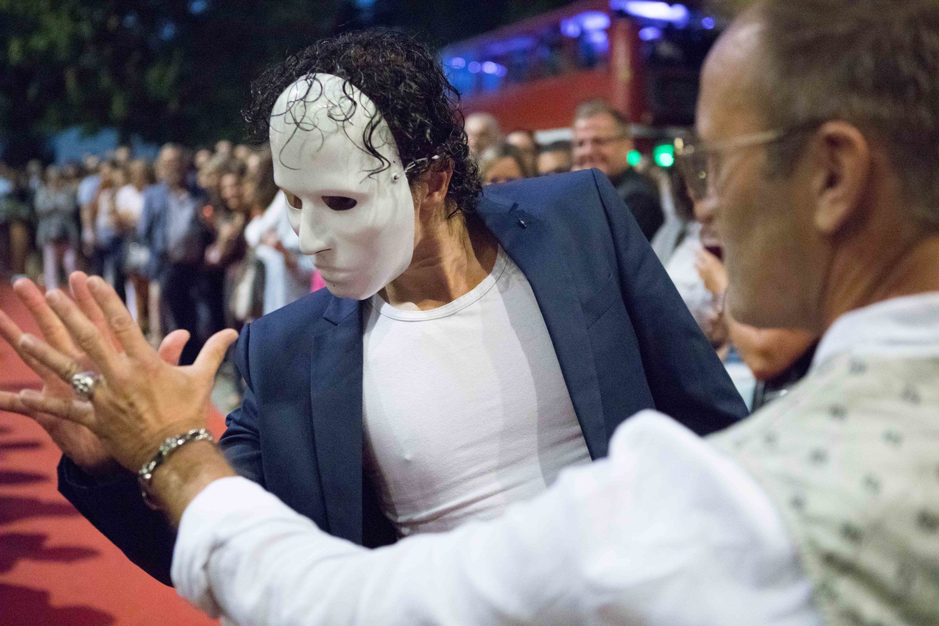 Ein Mann mit einer Maske tanzt mit einem anderen Mann auf einem roten Teppich.
