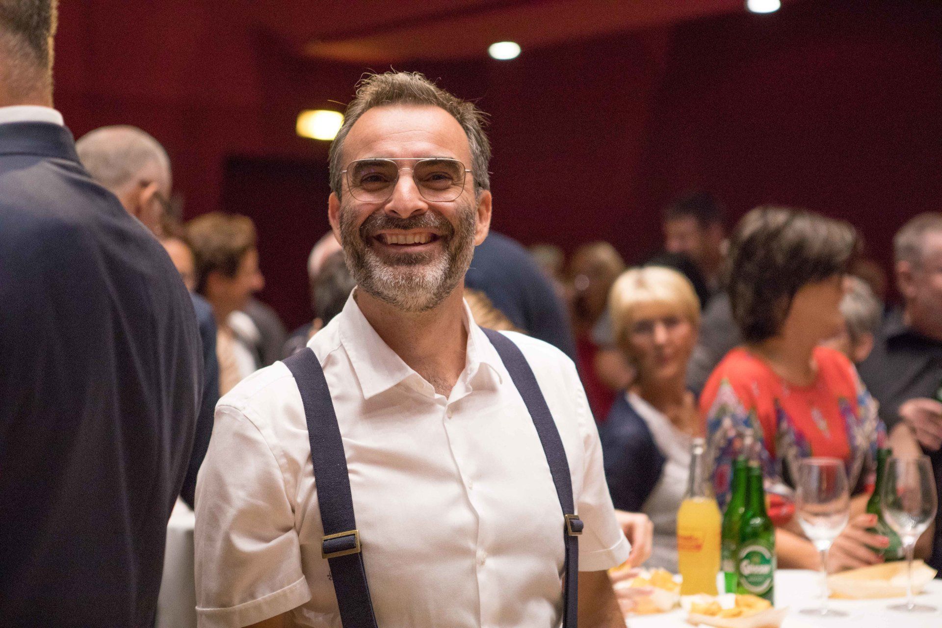 Ein Mann mit Bart und Hosenträgern lächelt in einem überfüllten Raum.