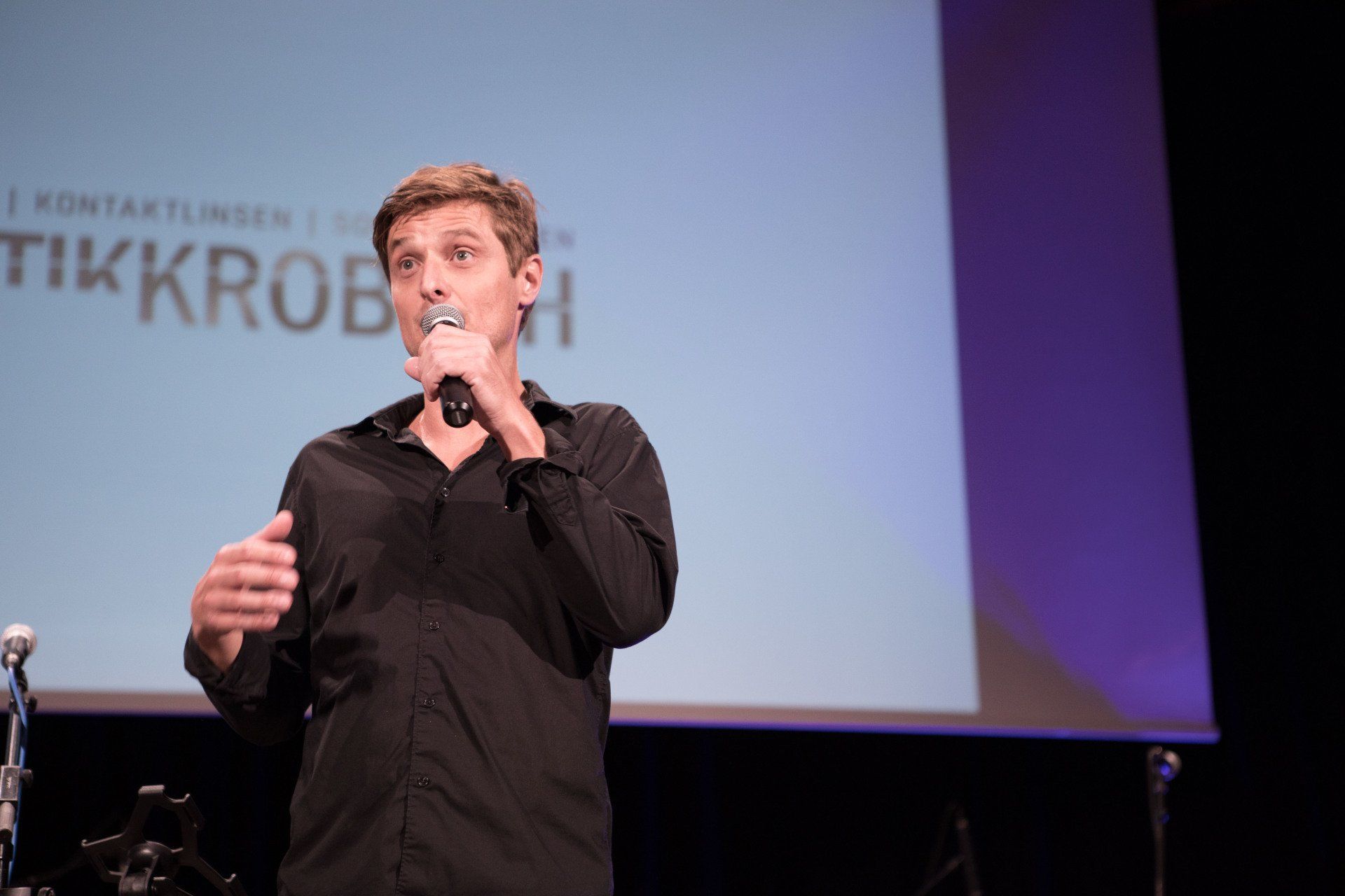 Ein Mann in einem schwarzen Hemd spricht auf der Bühne in ein Mikrofon.