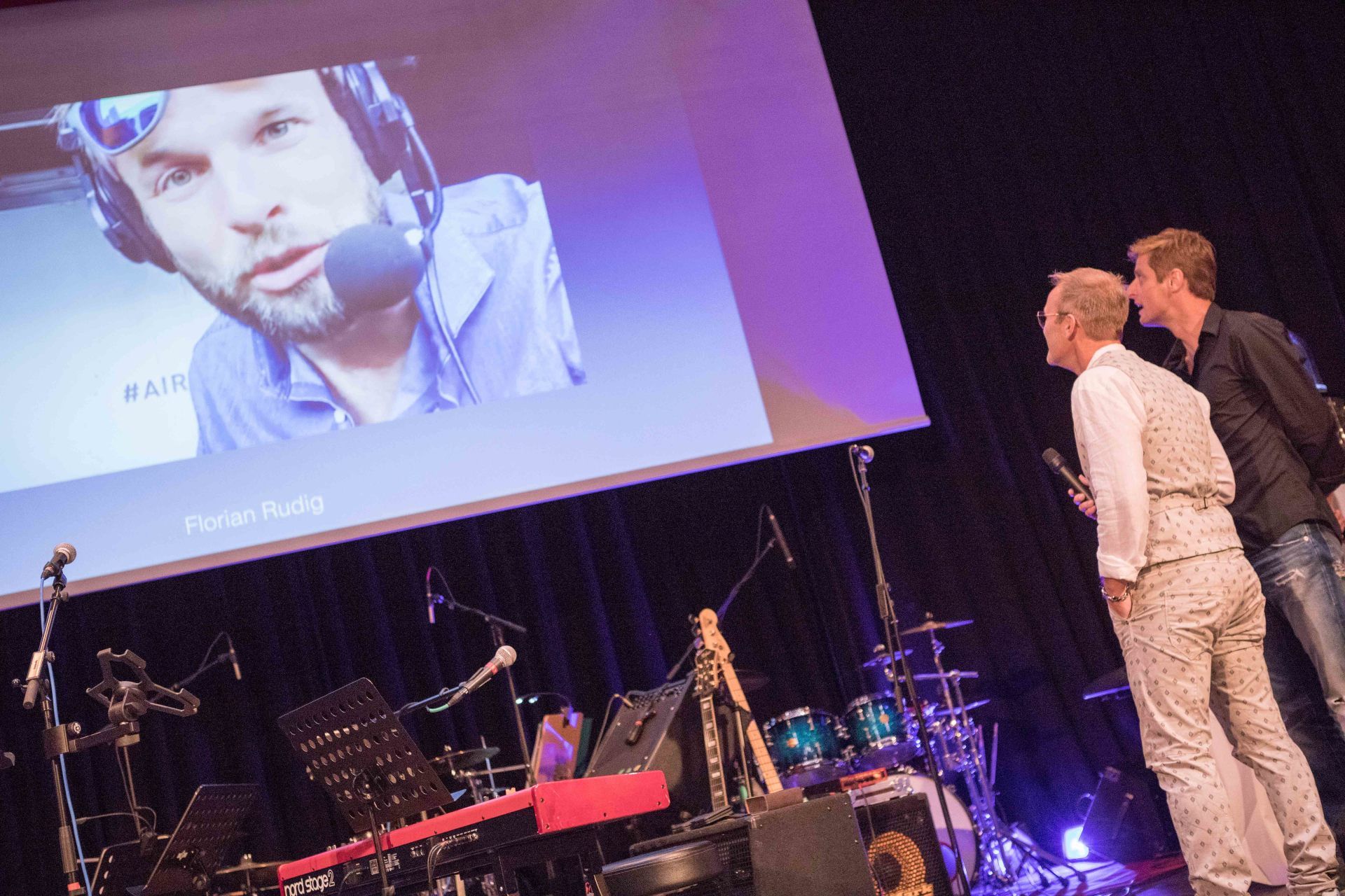 Zwei Männer stehen vor einem großen Bildschirm, auf dem das Bild eines Mannes zu sehen ist.