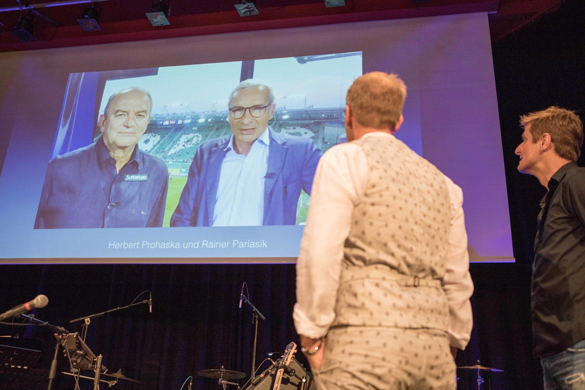 Zwei Männer stehen vor einem großen Bildschirm, auf dem drei Männer abgebildet sind.
