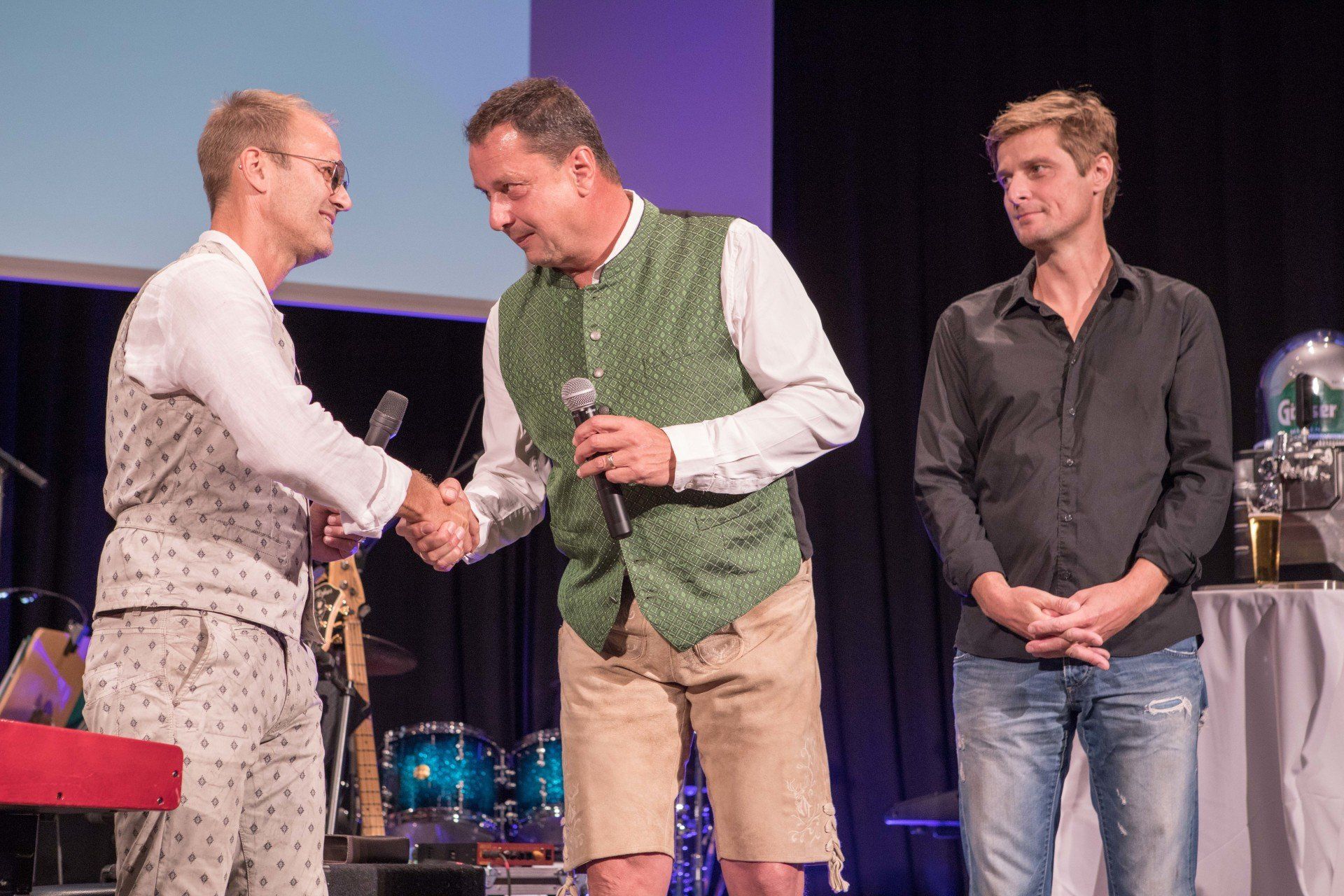 Ein Mann in einer grünen Weste schüttelt einem anderen Mann auf einer Bühne die Hand.