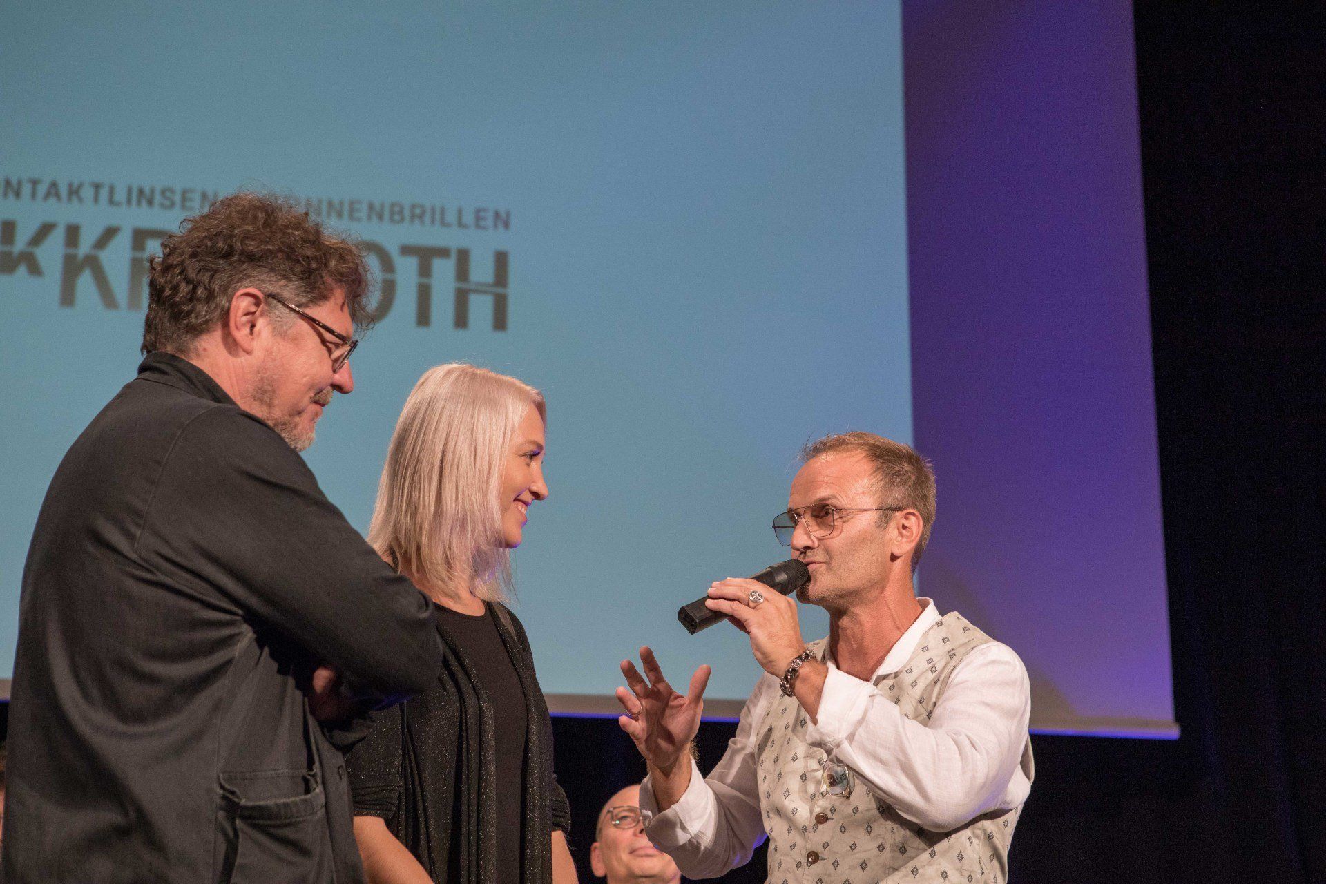 Ein Mann hält ein Mikrofon und spricht mit zwei Personen auf einer Bühne.