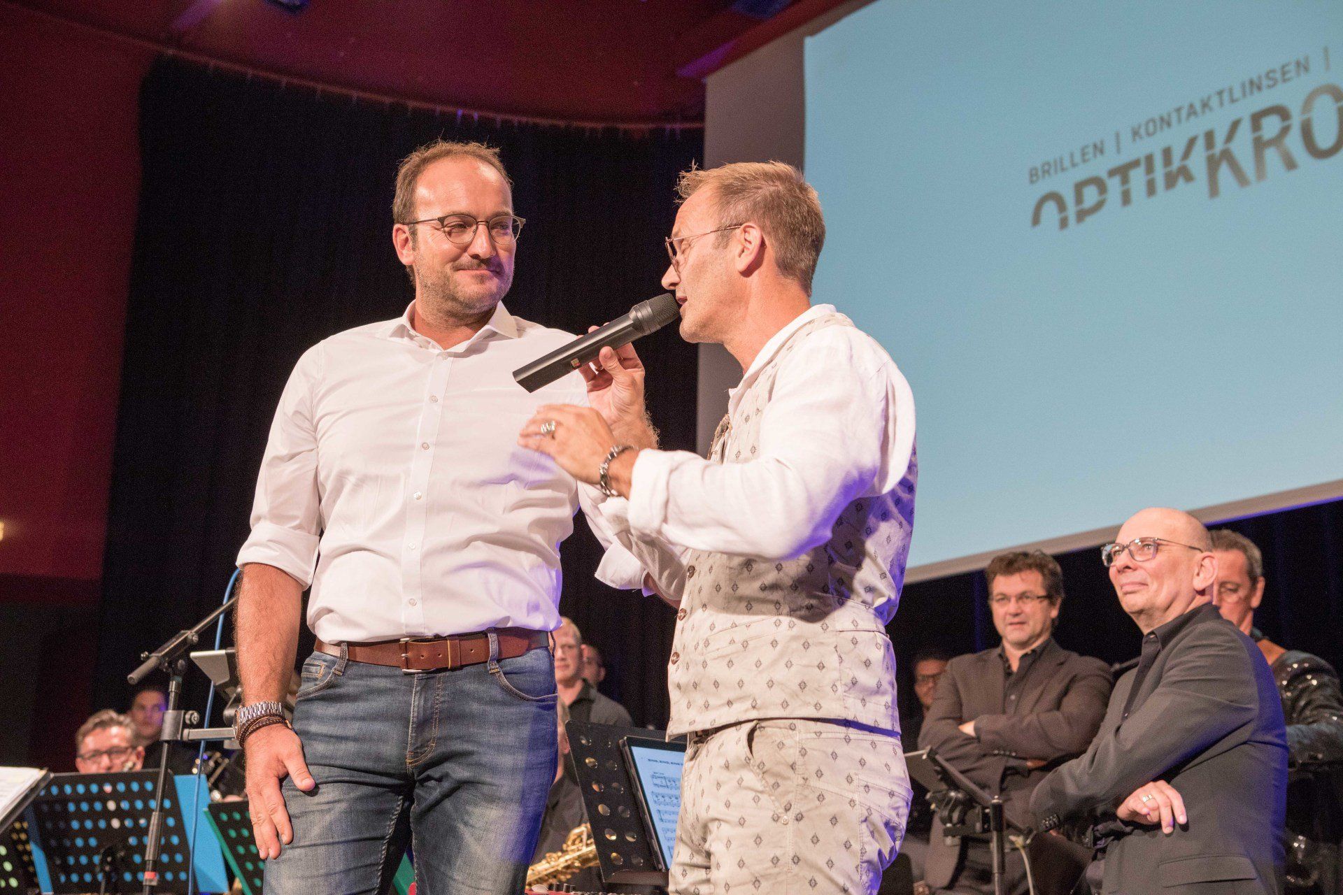 Zwei Männer stehen nebeneinander auf einer Bühne und einer hält ein Mikrofon.