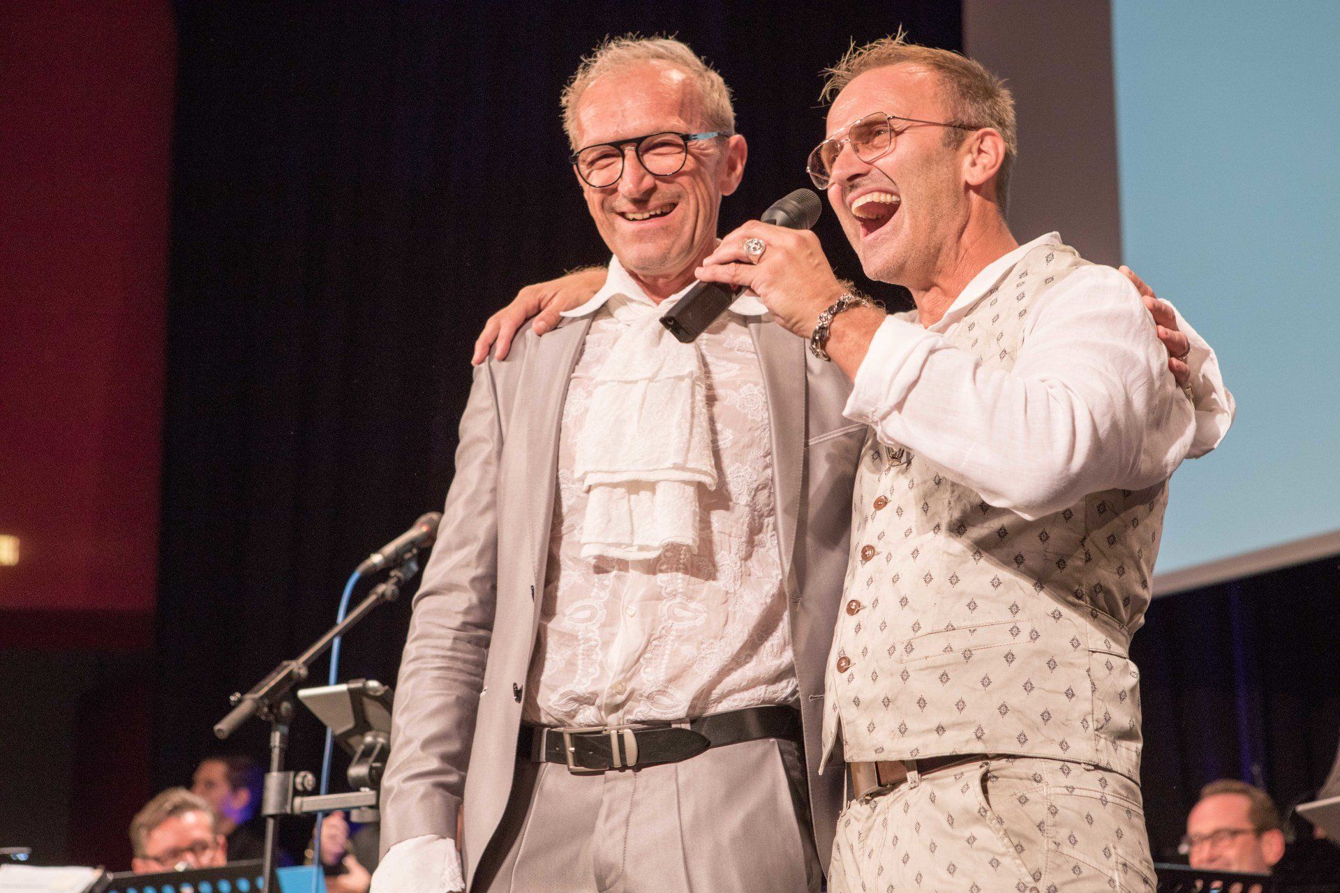 Zwei Männer stehen nebeneinander auf einer Bühne und einer singt in ein Mikrofon.