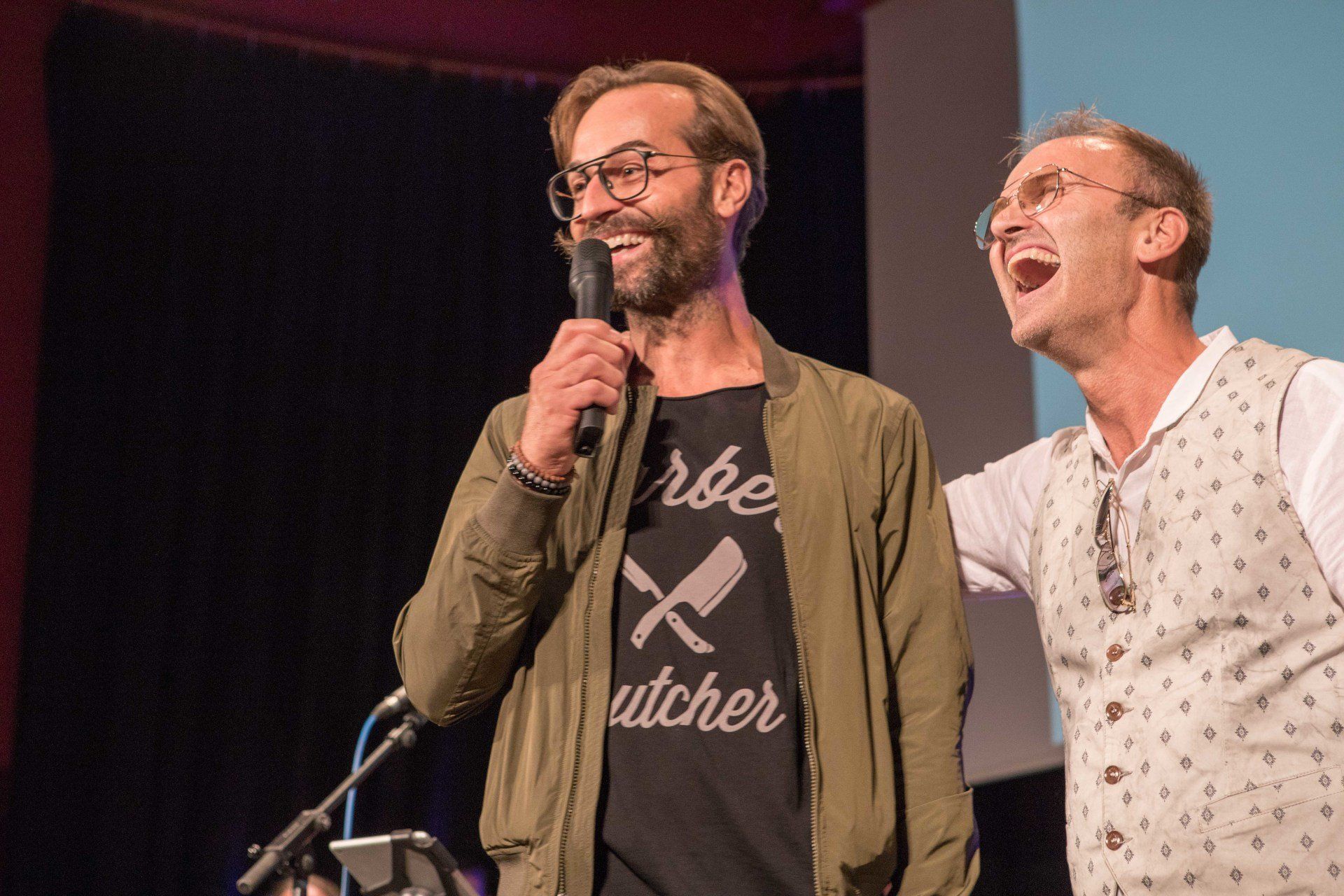 Zwei Männer stehen nebeneinander auf einer Bühne und einer von ihnen hält ein Mikrofon.