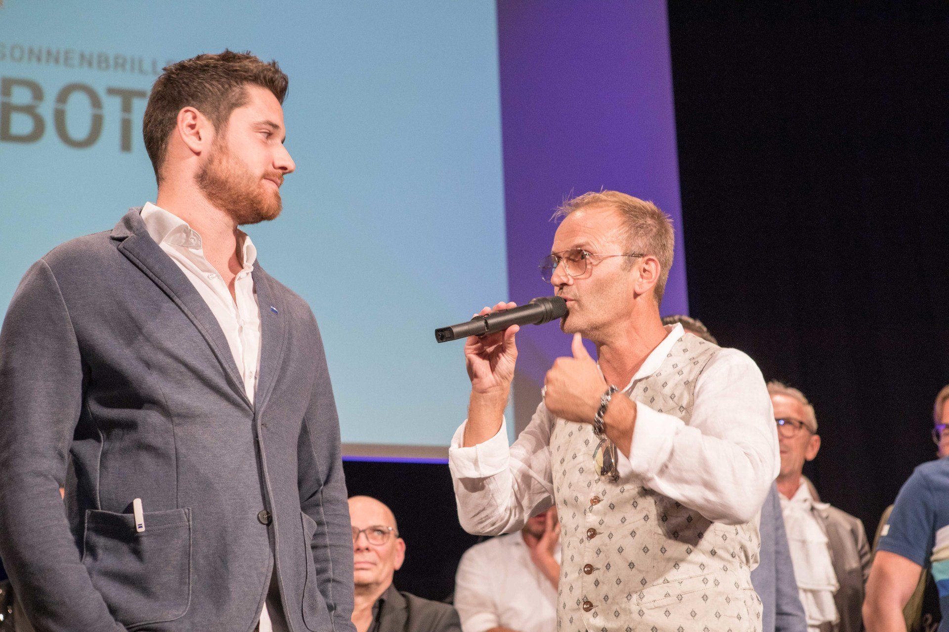 Zwei Männer stehen nebeneinander auf einer Bühne und sprechen in Mikrofone.