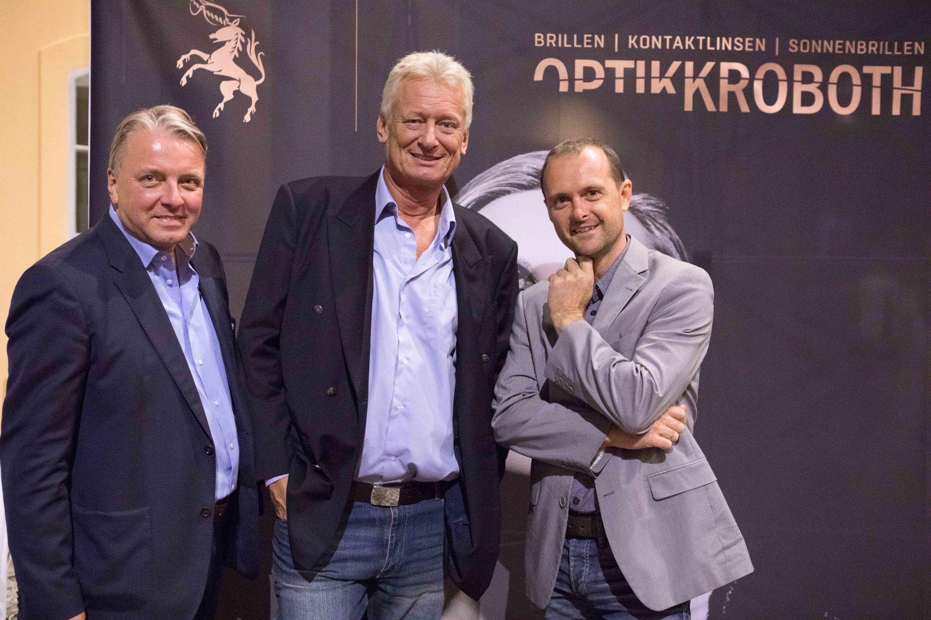 Drei Männer posieren für ein Foto vor einem Schild mit der Aufschrift „Optikkrobotn“