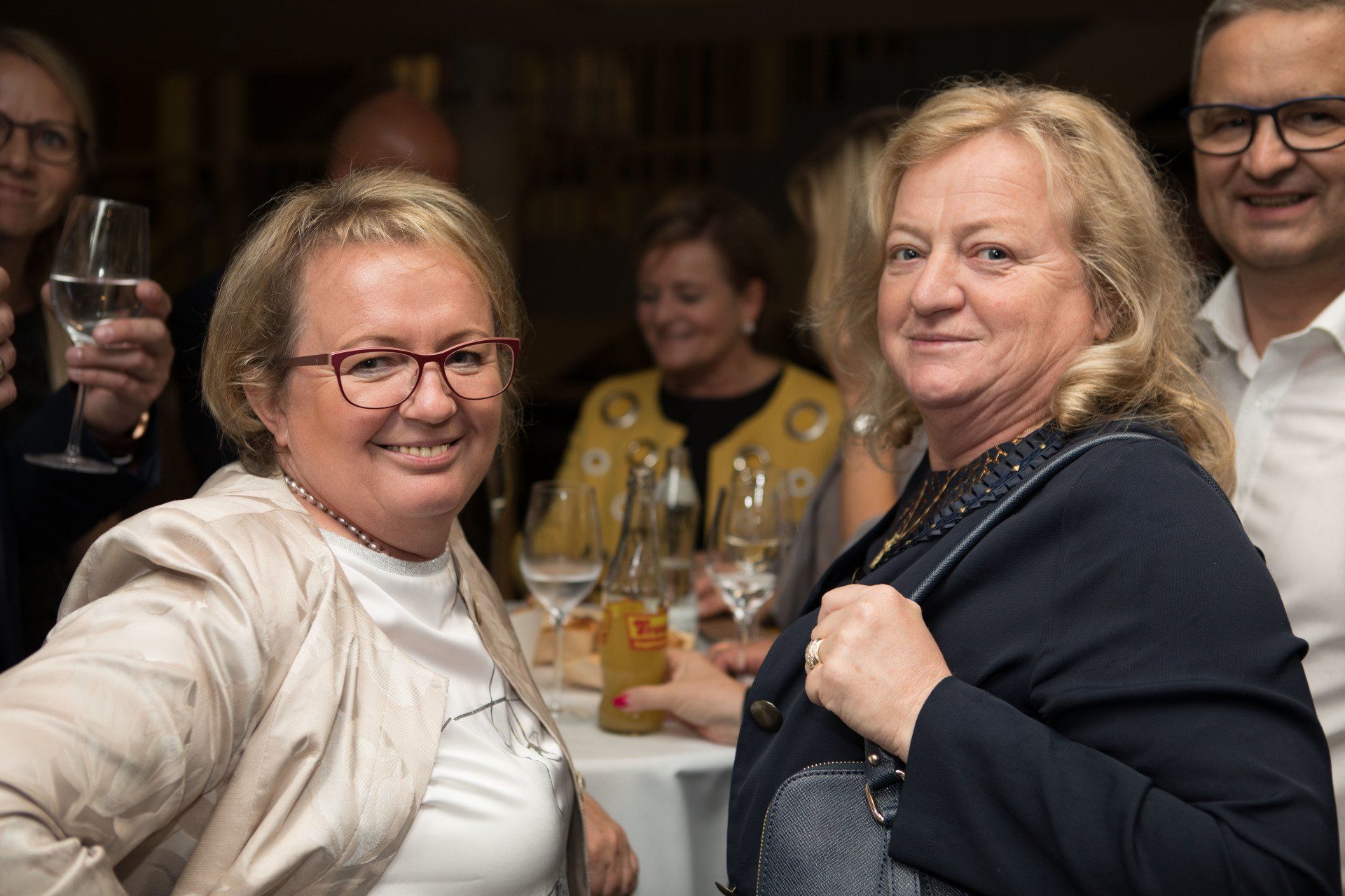 Zwei Frauen posieren für ein Foto, während sie mit Weingläsern an einem Tisch sitzen.