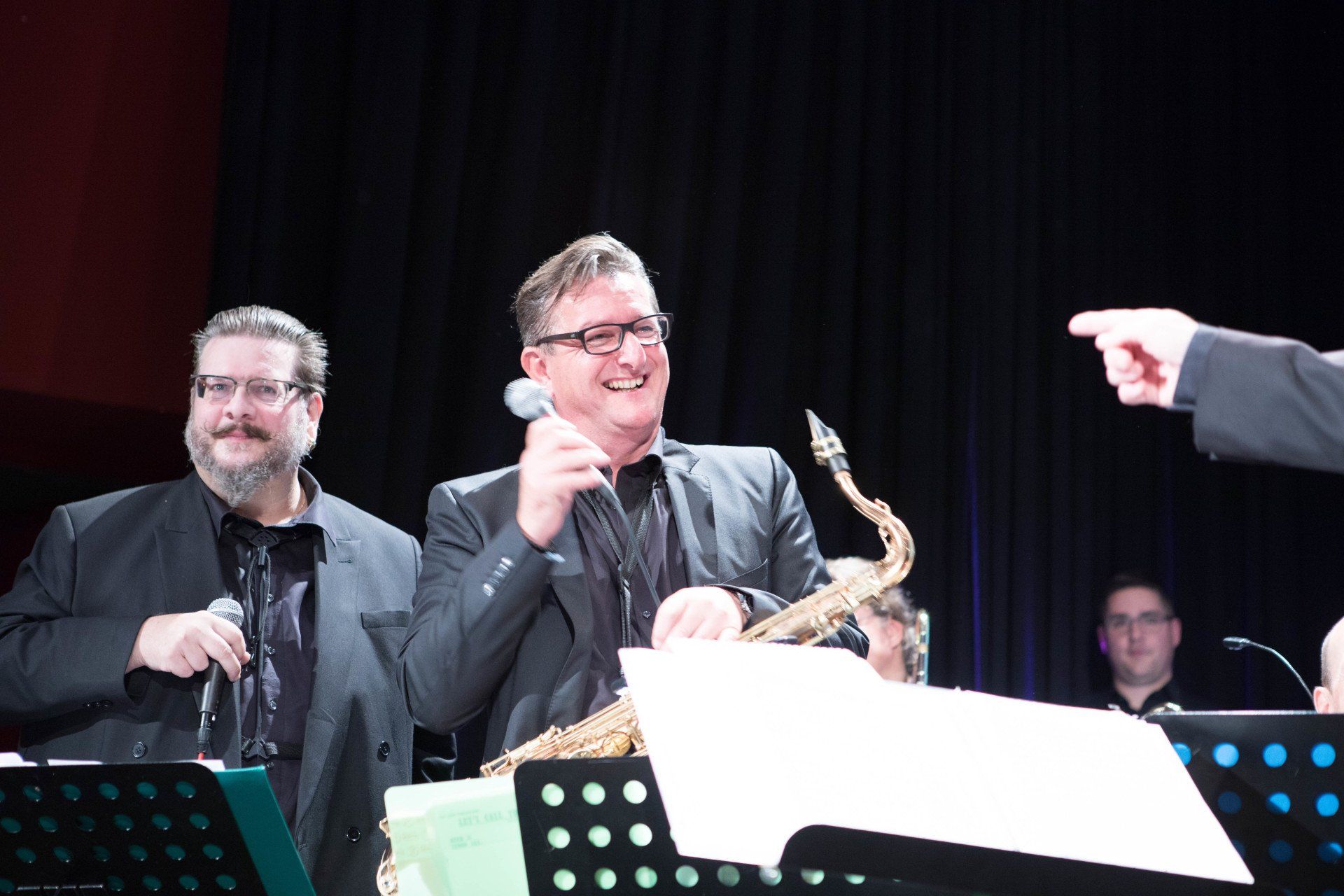 Zwei Männer stehen mit einem Saxophon und einem Mikrofon nebeneinander auf einer Bühne.