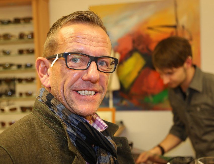 Ein Mann mit Brille und Schal lächelt vor einem Gemälde.