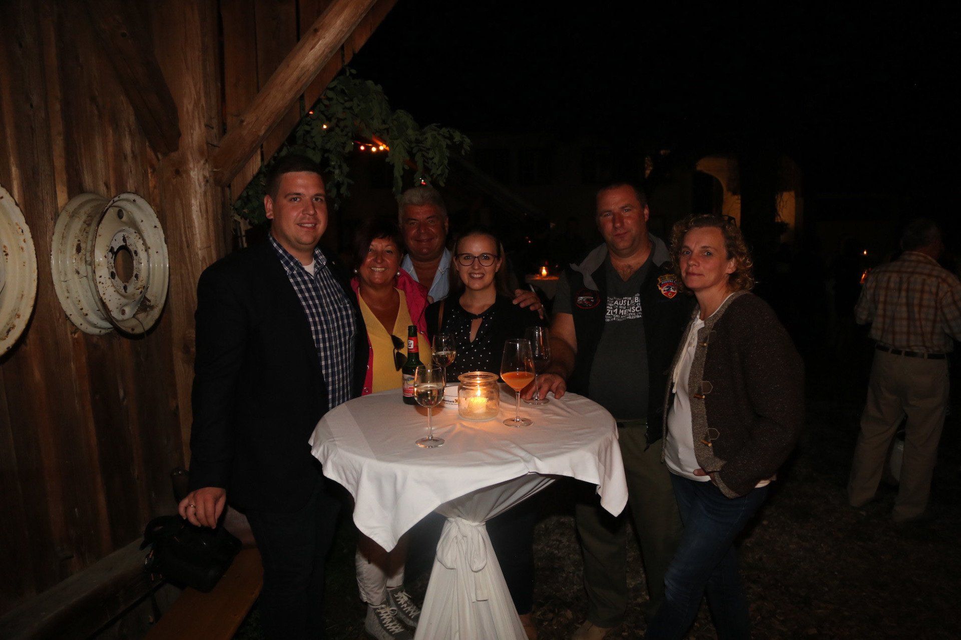 Eine Gruppe von Menschen steht mit Weingläsern und einer Kerze um einen Tisch herum.