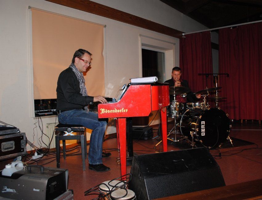 Ein Mann spielt ein rotes Klavier, während ein anderer Mann Schlagzeug spielt