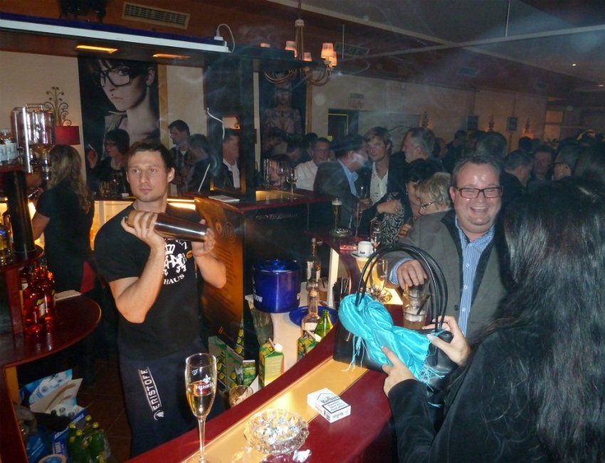 Eine Gruppe von Menschen hat sich in einer Bar versammelt, darunter ein Mann, der ein schwarzes T-Shirt mit der Aufschrift „Just do it“ trägt.