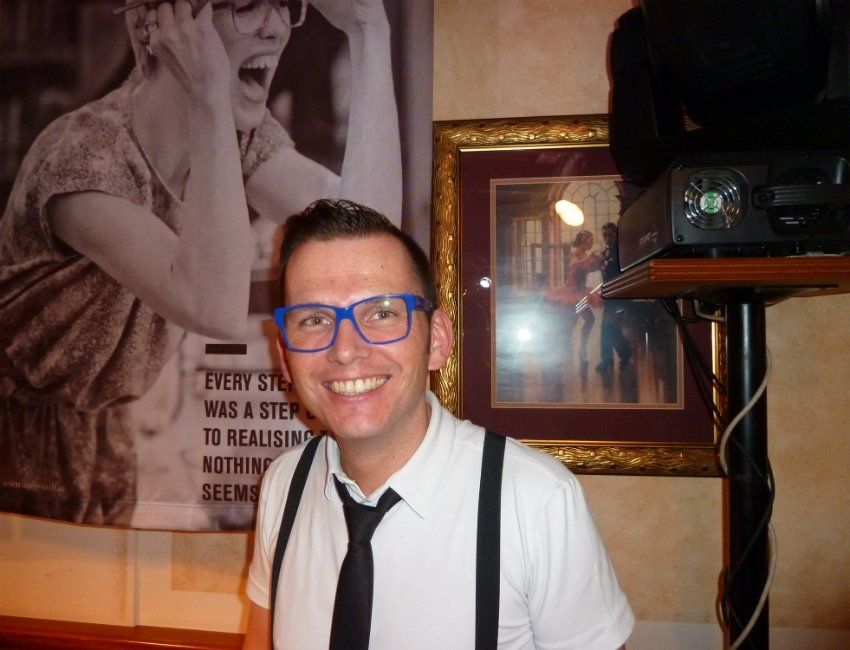 Ein Mann mit blauer Brille und Hosenträgern lächelt vor einem Plakat mit der Aufschrift „Jeder Schritt war ein Schritt“.