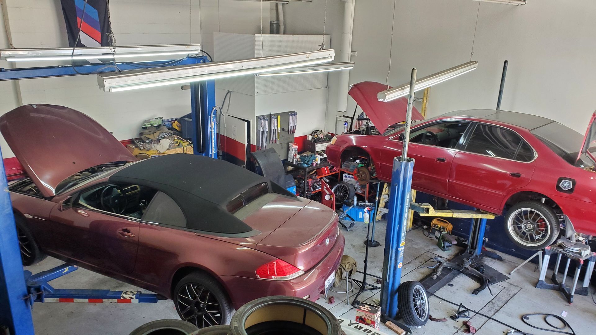 Three cars in a cluttered garage, including a maroon convertible and a red coupe on lifts