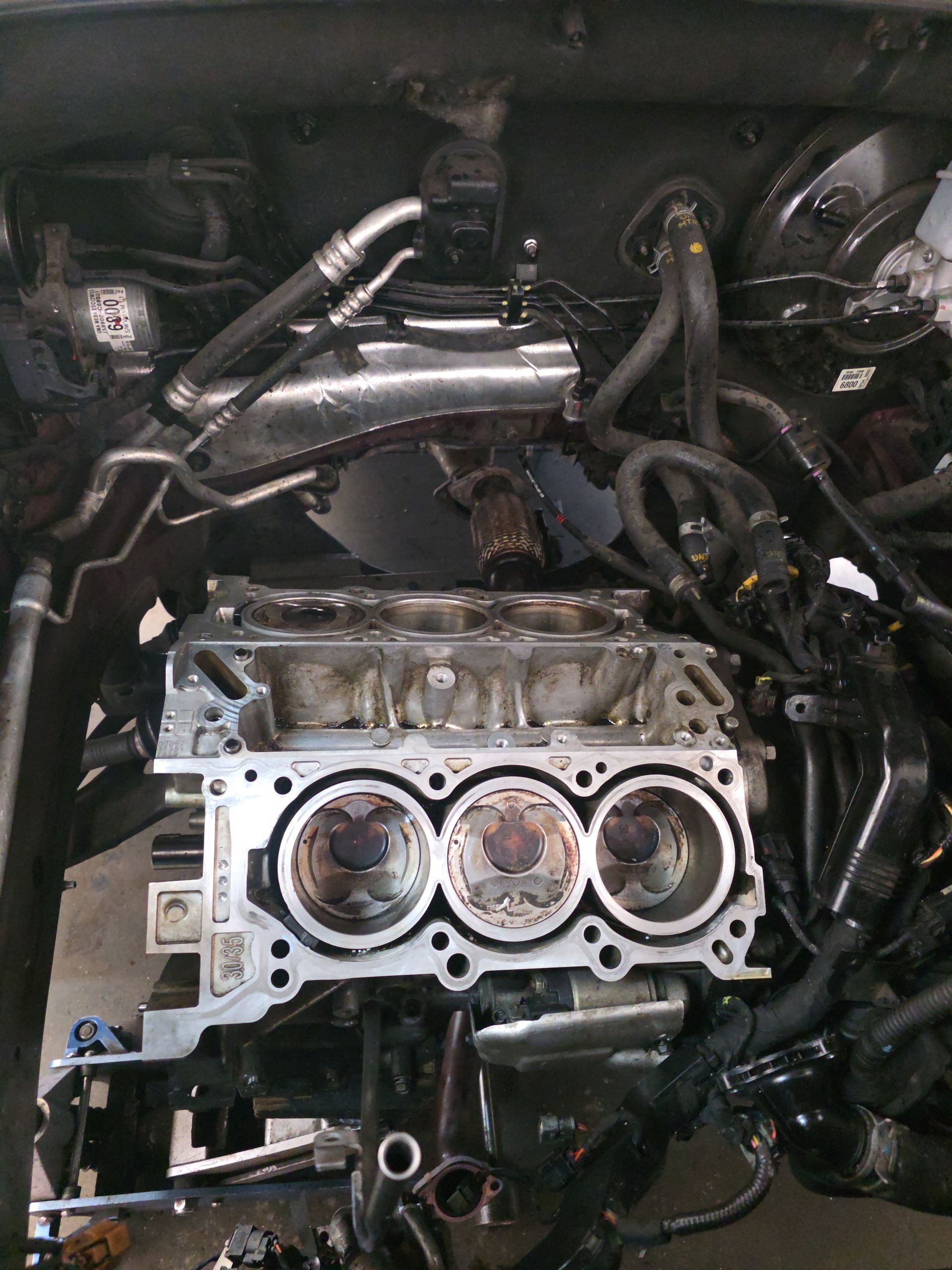 Car engine bay with cylinder head exposed and removed, showing three visible piston tops and hoses around it