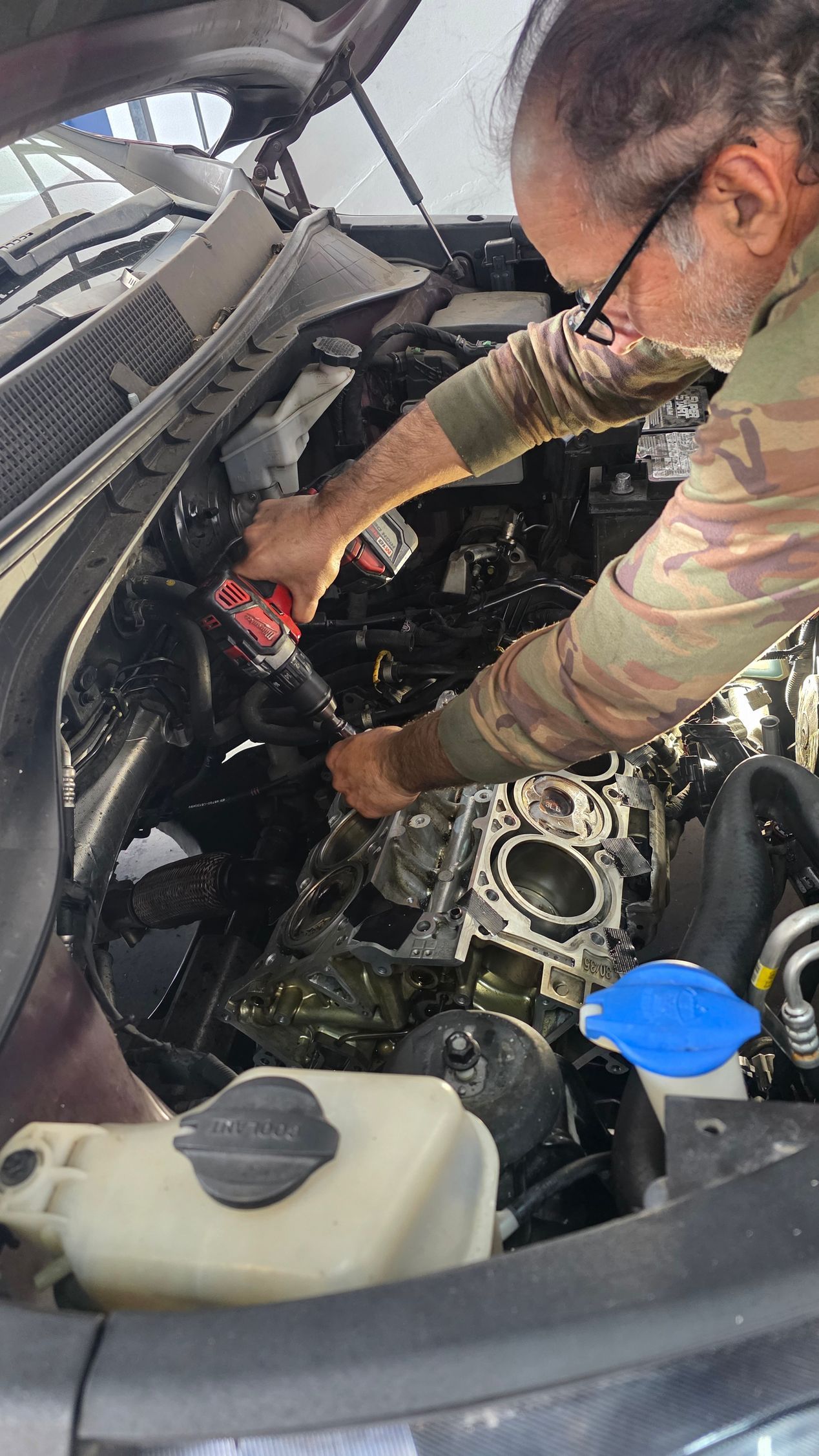 Mechanic working under a car hood, inspecting engine components near a blue fluid cap.