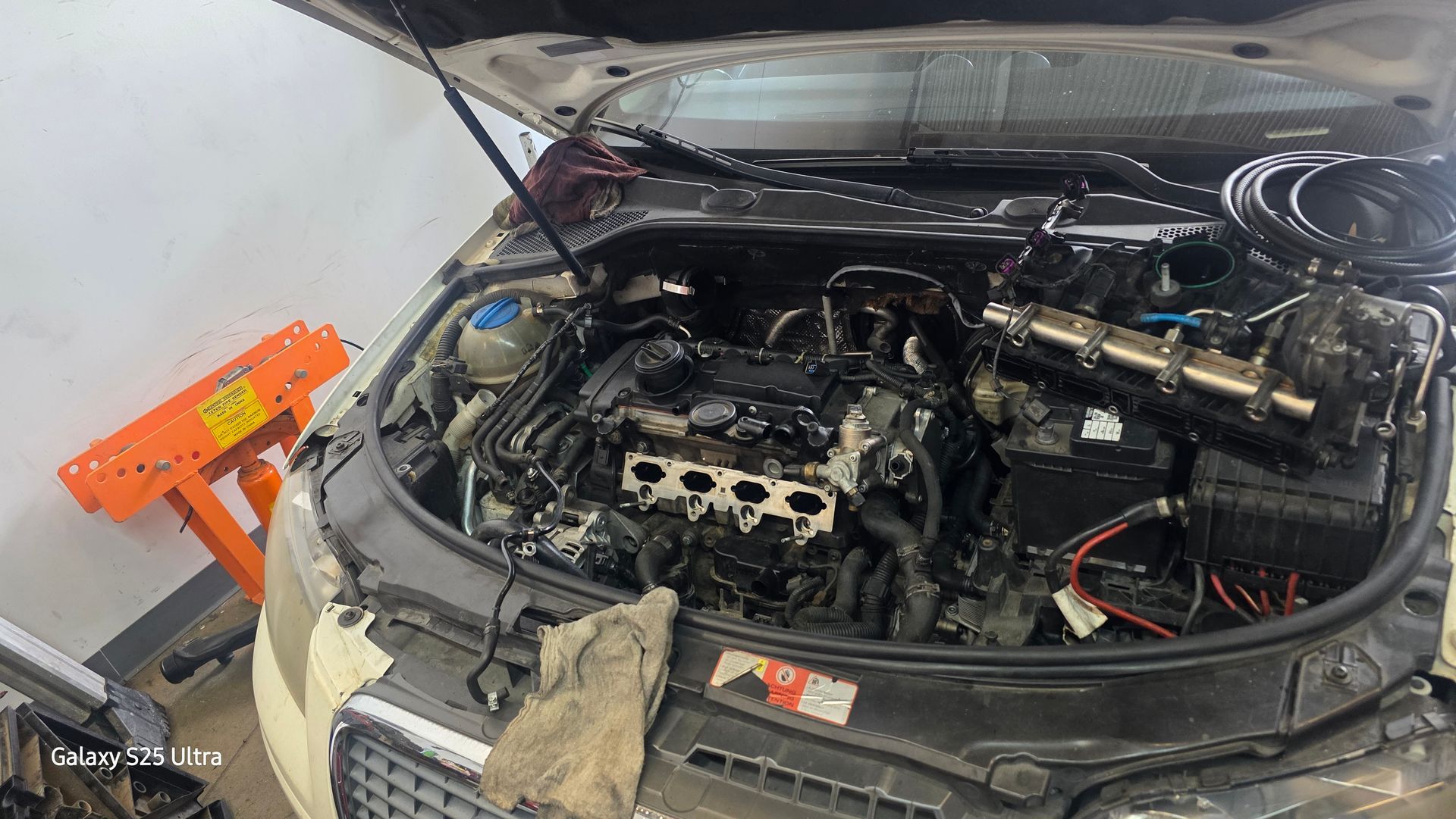 Car engine bay with hood open, engine partly disassembled in a garage.