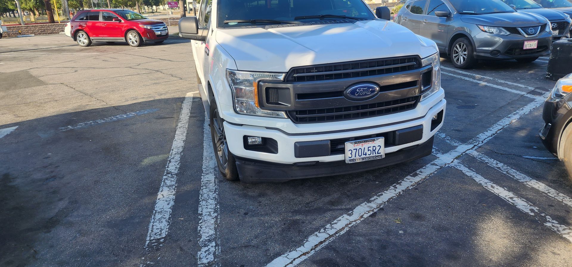 White Ford truck parked front-in across two spaces in a parking lot, with other cars nearby.