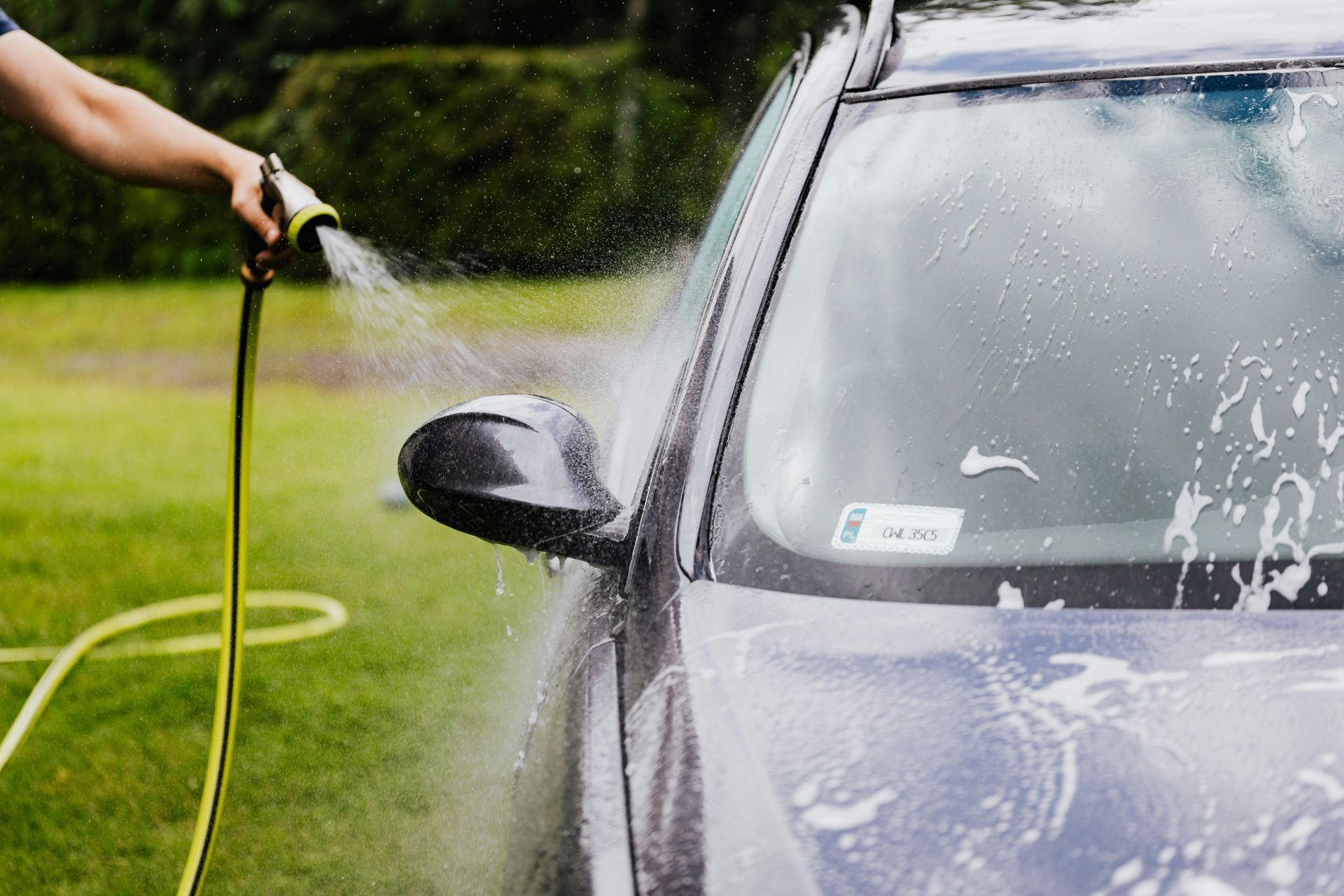 A person washing a dark blue car with a hose in a grassy area.