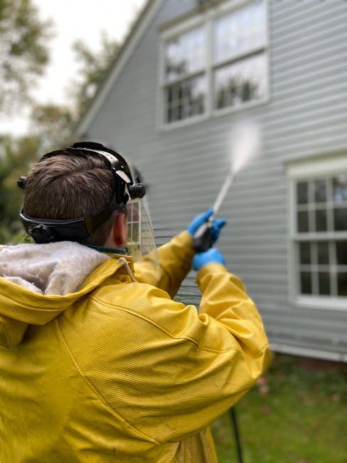 Person in yellow rain gear power washing a gray house.