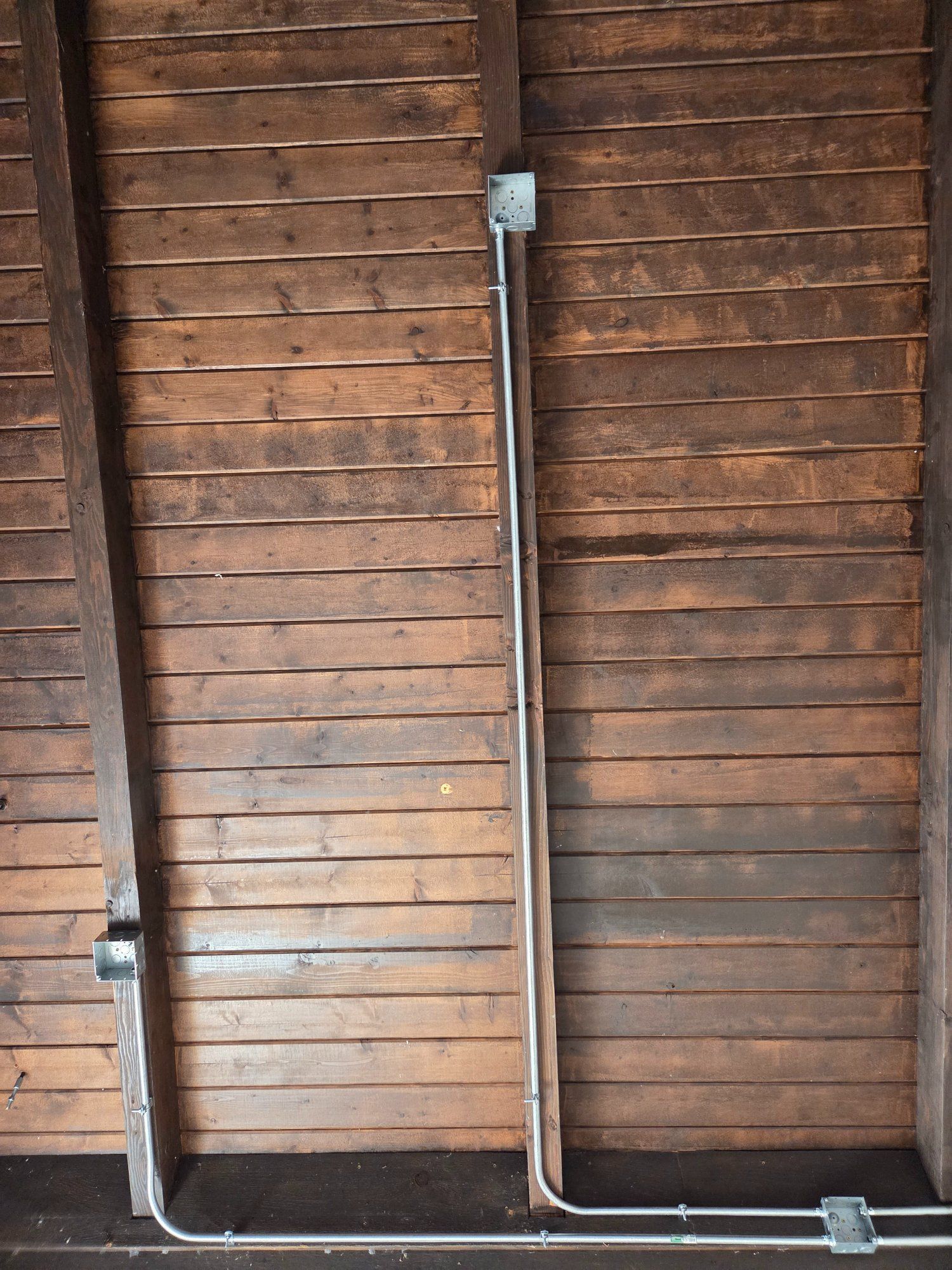 Metal electrical conduit runs vertically on weathered wooden planks, connected to boxes at top and bottom.