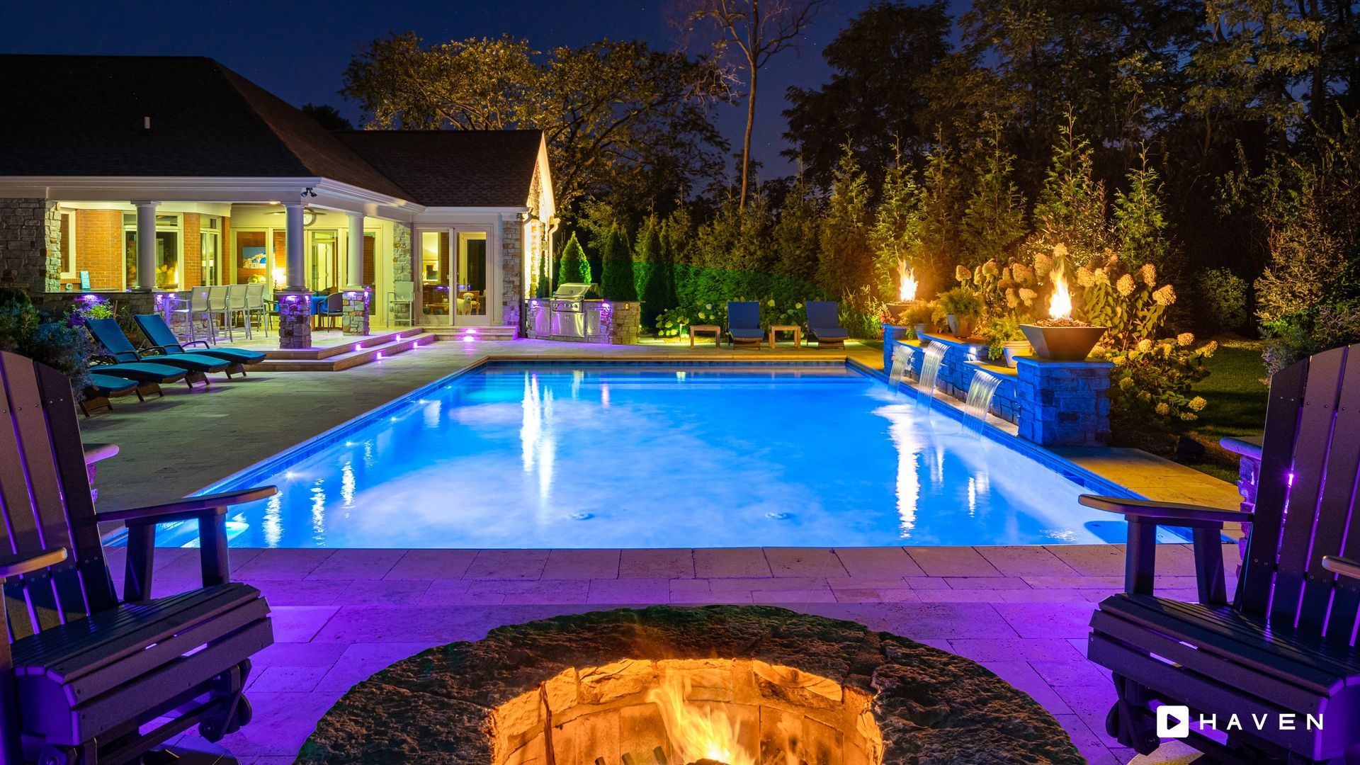 A large swimming pool is lit up at night with a fire pit in the middle.