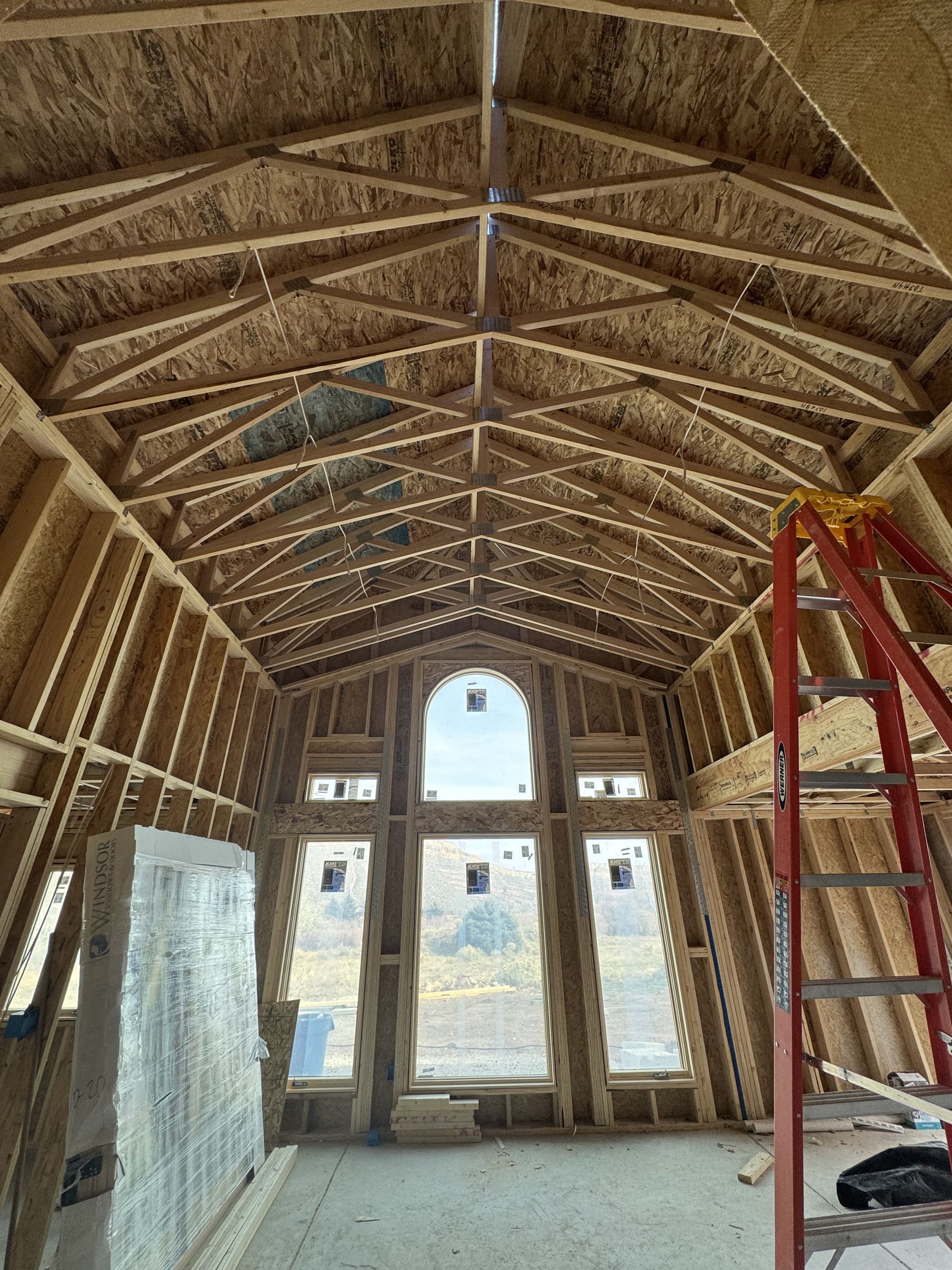 Interior of a wooden house under construction, with high arched ceiling and large windows. A ladder stands at the side.