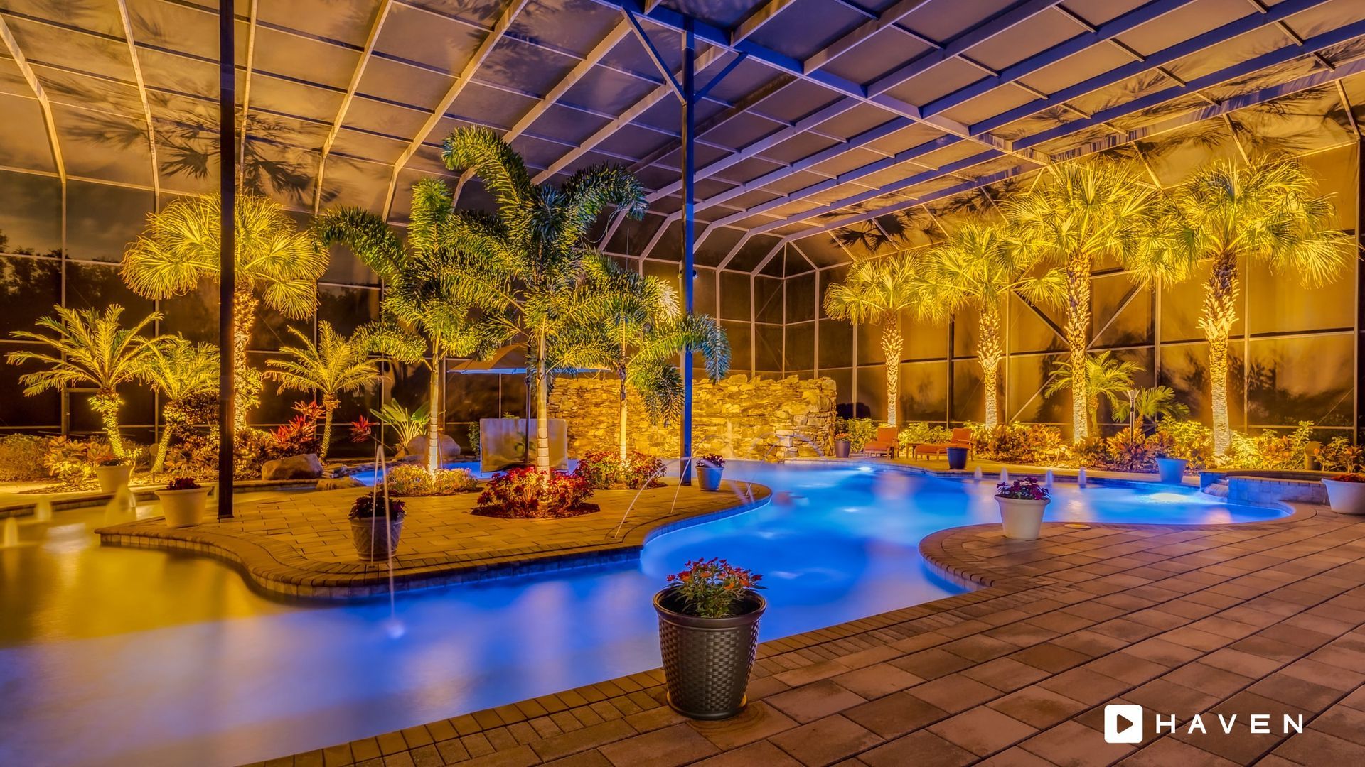 A large swimming pool is lit up at night with palm trees in the background.
