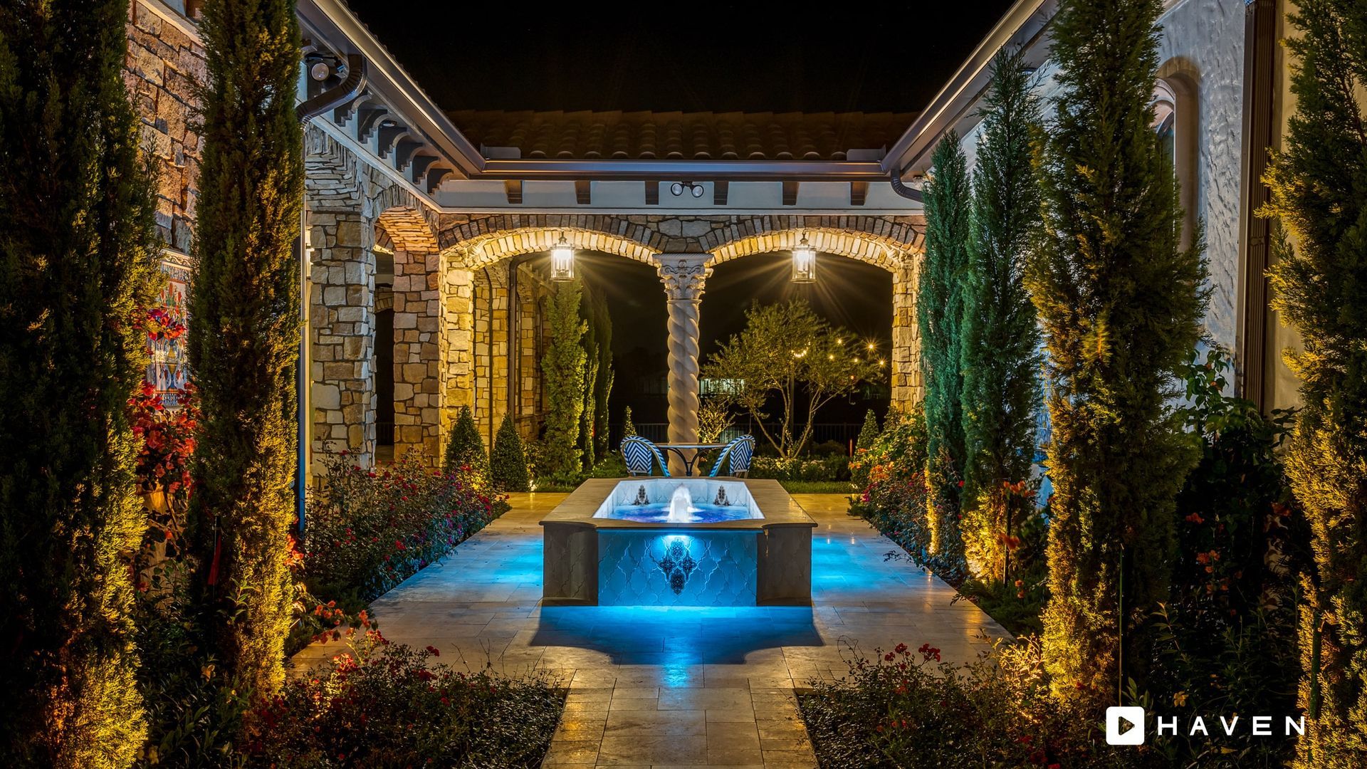 A luxury outdoor courtyard at night, lit by blue and yellow lights. Features a fountain and stone arches.