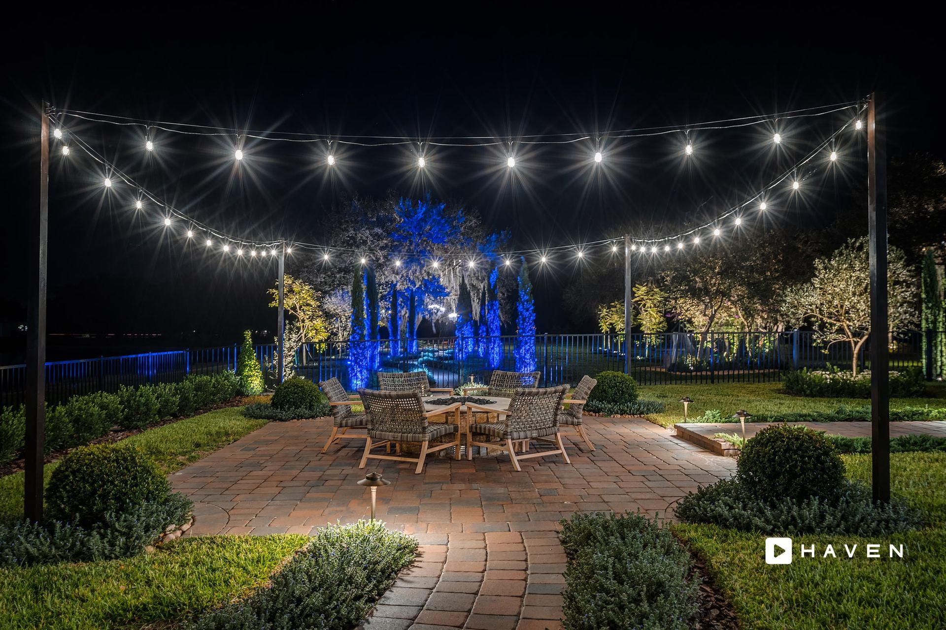 Nighttime outdoor patio lit by string lights over a dining table and chairs. Green bushes and trees surround a brick patio.