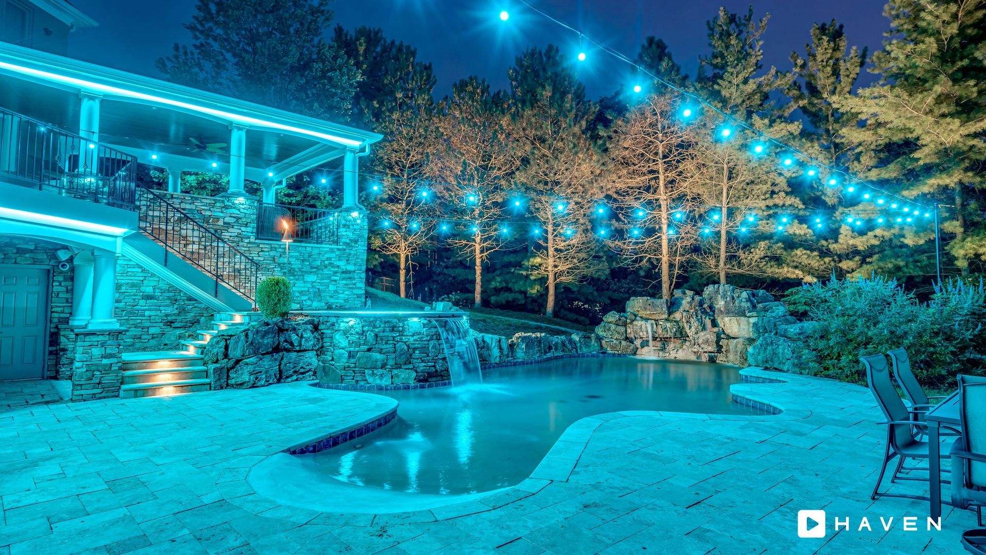 A large swimming pool is surrounded by trees and lights at night.