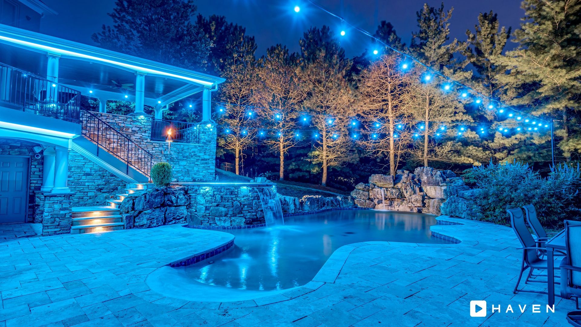 A large swimming pool is surrounded by trees and lights at night.