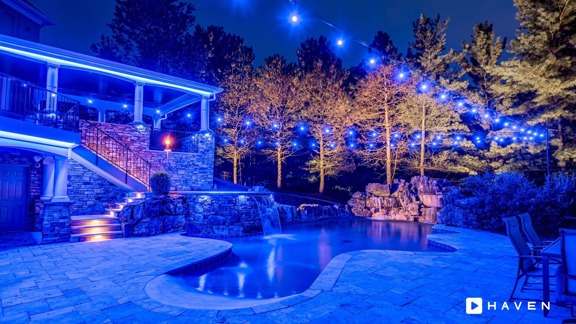 A backyard with a pool and trees lit up with blue lights at night.