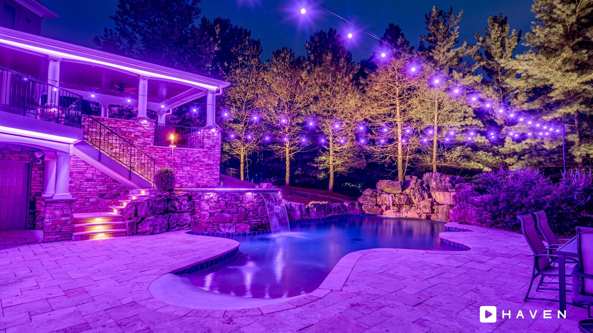 A swimming pool is lit up with purple lights in the backyard of a house.