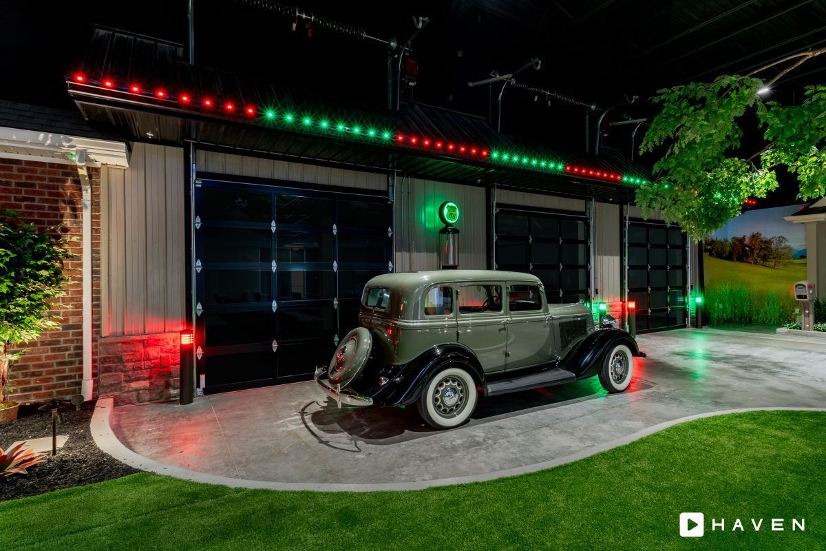An old car is parked in front of a garage with christmas lights.
