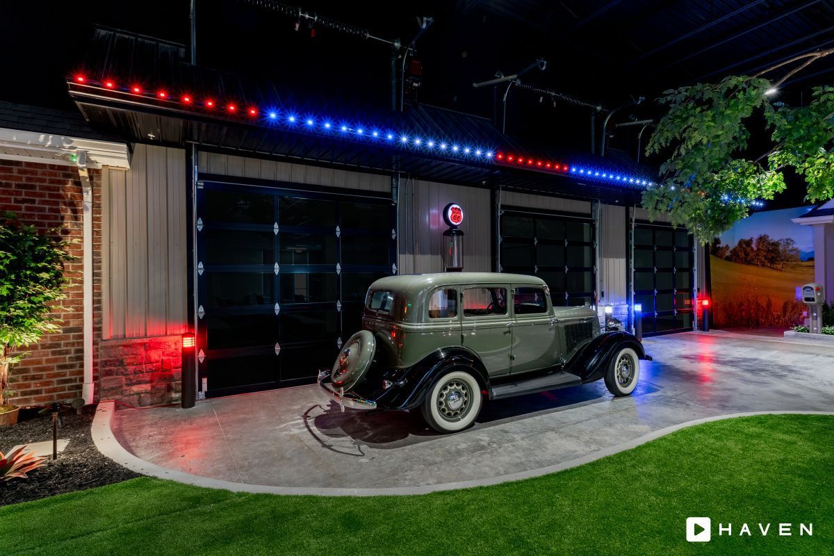 An old car is parked in front of a garage with red , white and blue lights.