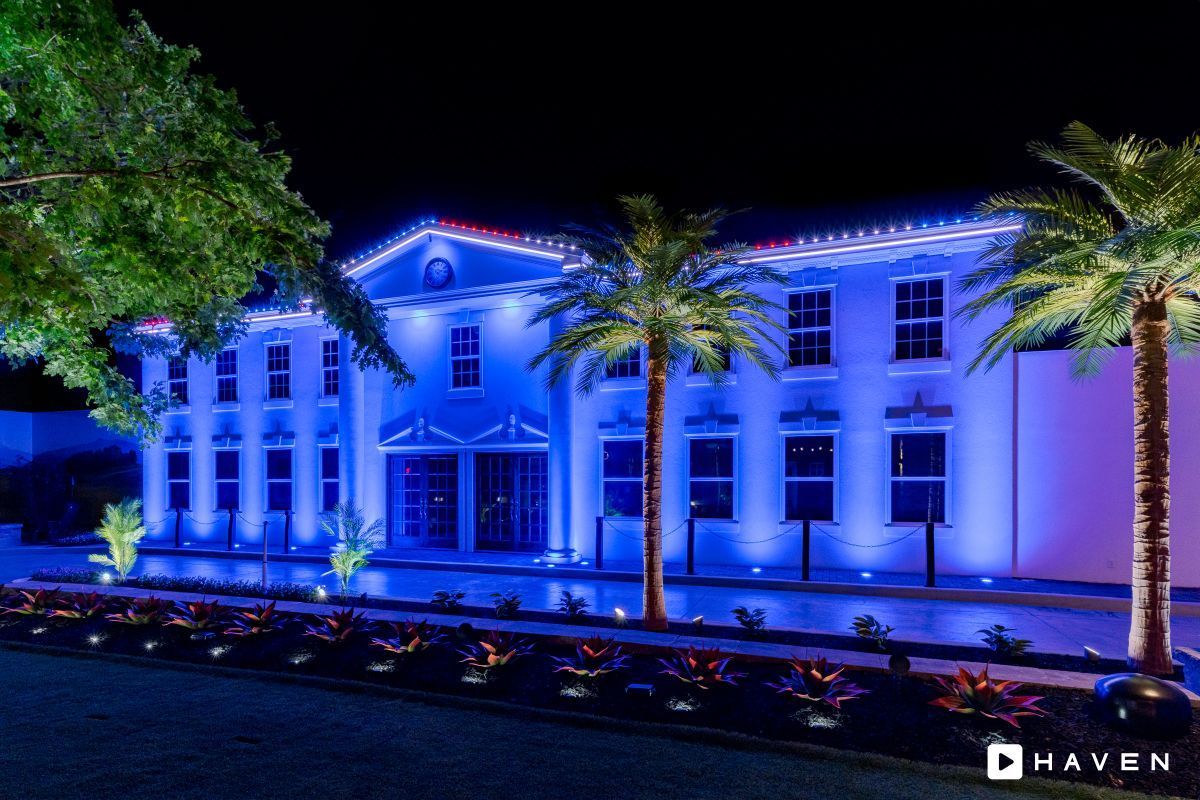 A blue-lit two-story building at night, with palm trees and landscaping. The building has a classical design.