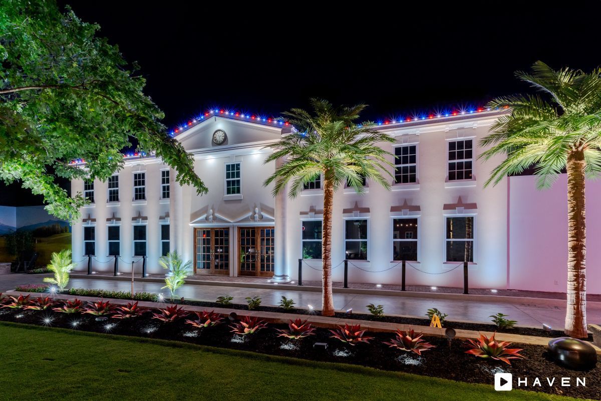Night view of a two-story white building with palm trees and red, white, and blue lights.