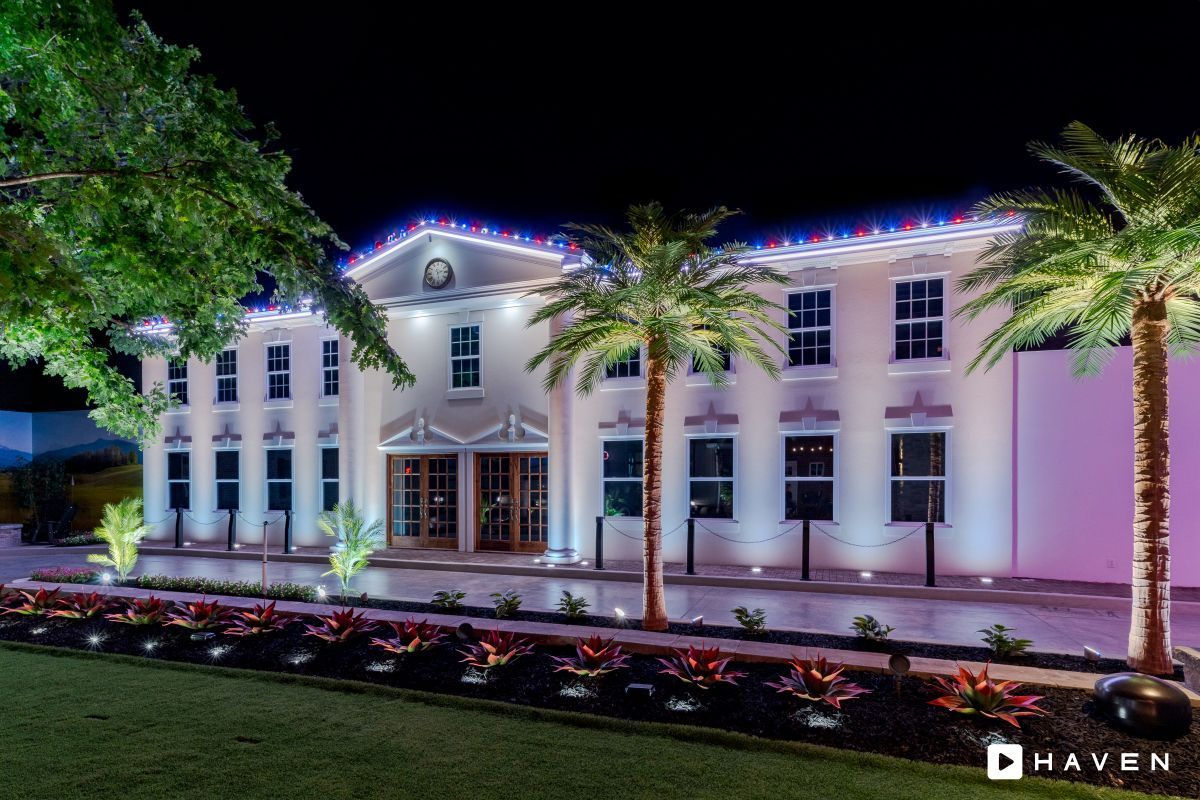 Elegant two-story building lit up at night with palm trees and manicured landscaping. Festive lights adorn the roofline.