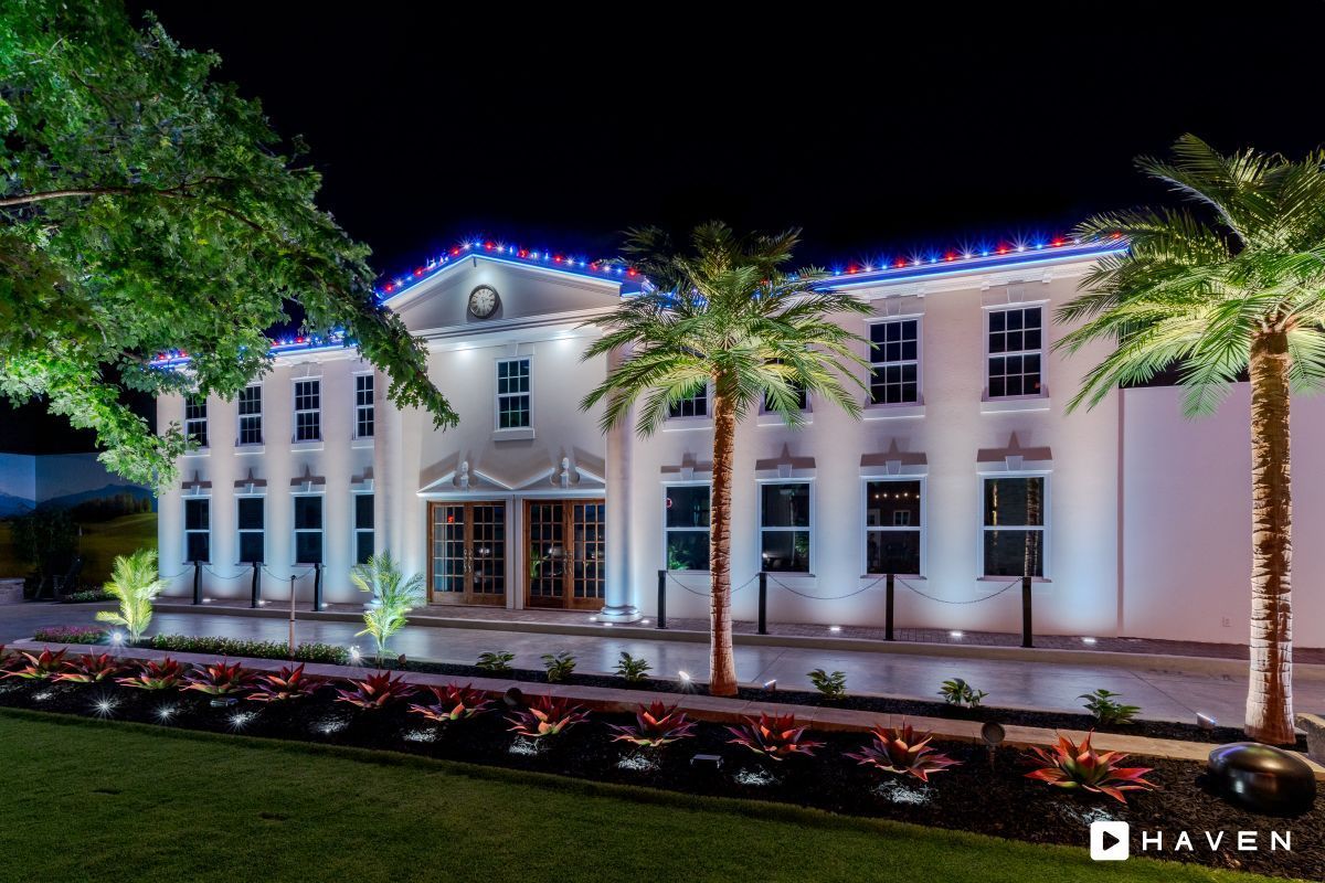 Night view of a two-story white building with palm trees and colorful lights on its roof and surrounding landscaping.