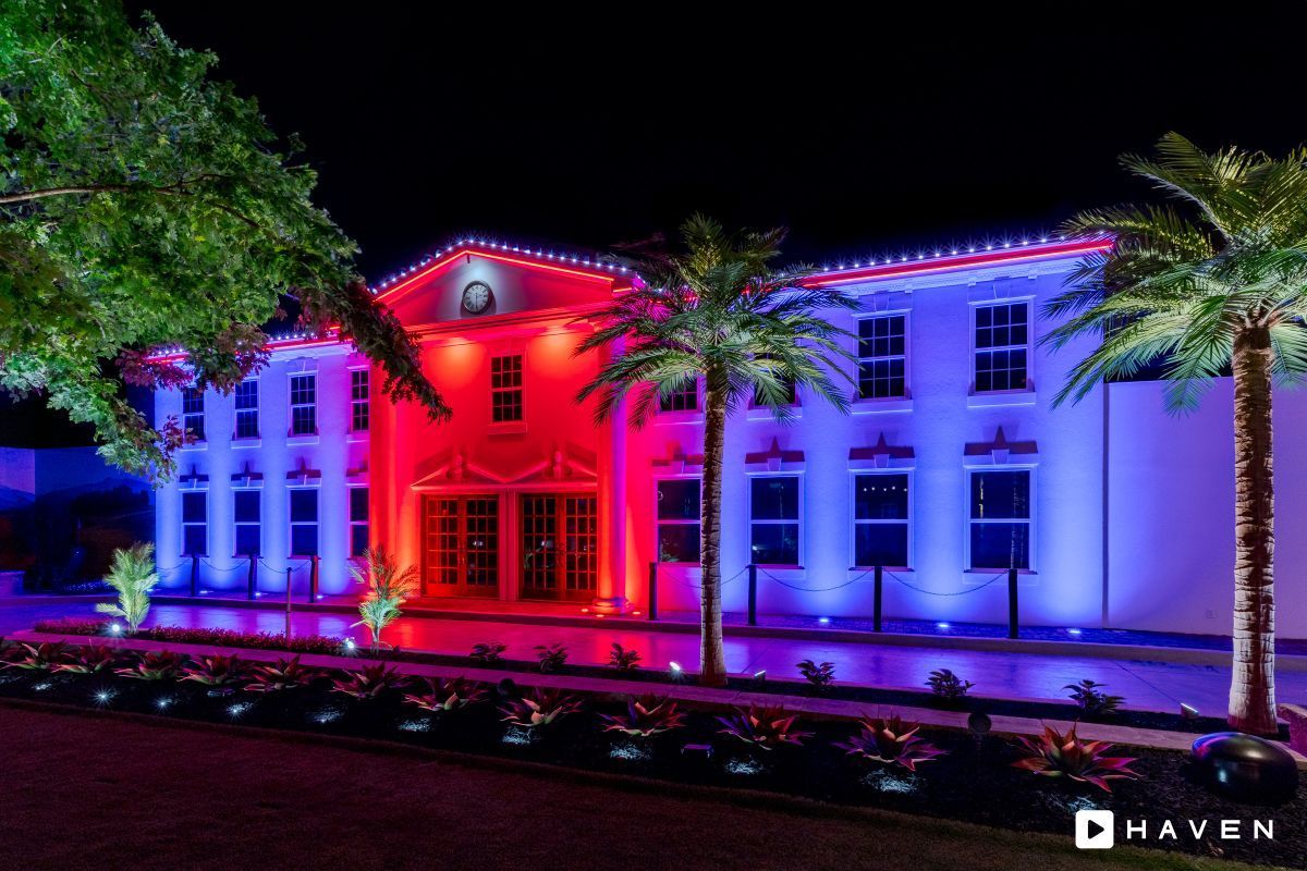A two-story building at night illuminated with red and blue lights, flanked by palm trees, in a landscaped setting.