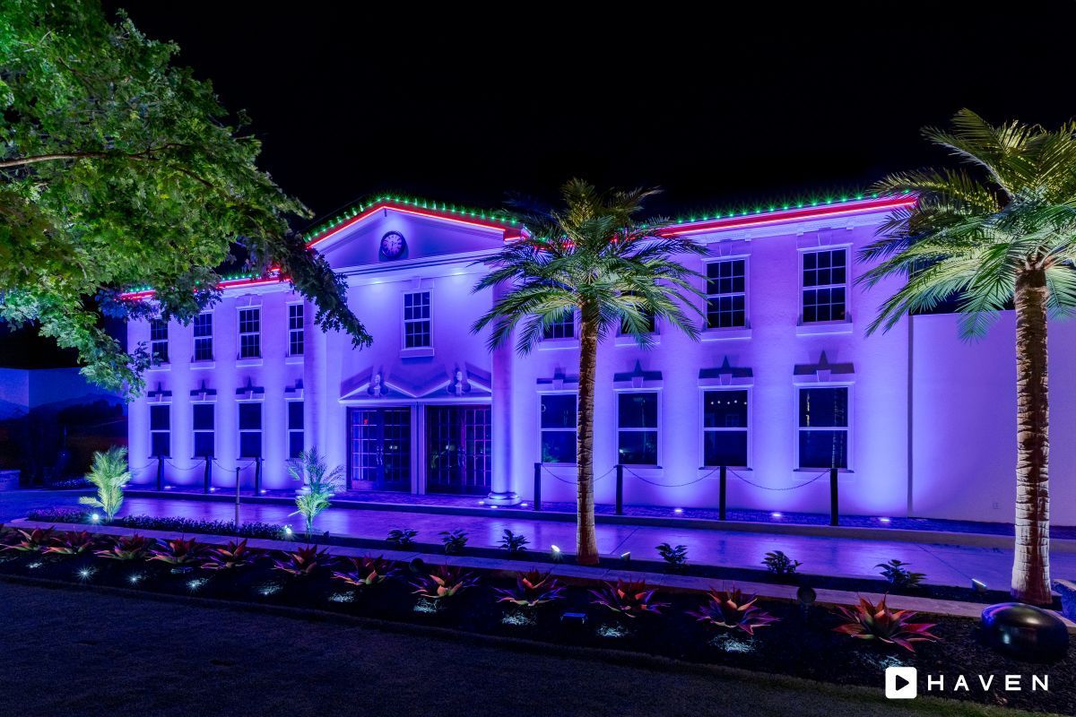 A large white building is lit up with purple lights and palm trees.