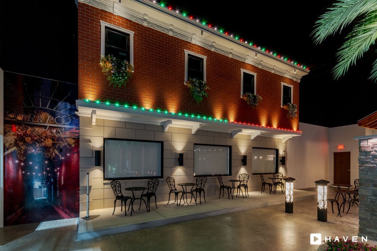 Exterior of a building at night with cafe tables and Christmas lights. A mural and palm tree are to the left.