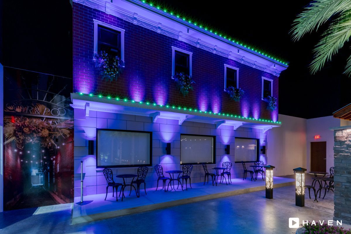 A building with tables and chairs in front of it is lit up with purple and green lights.