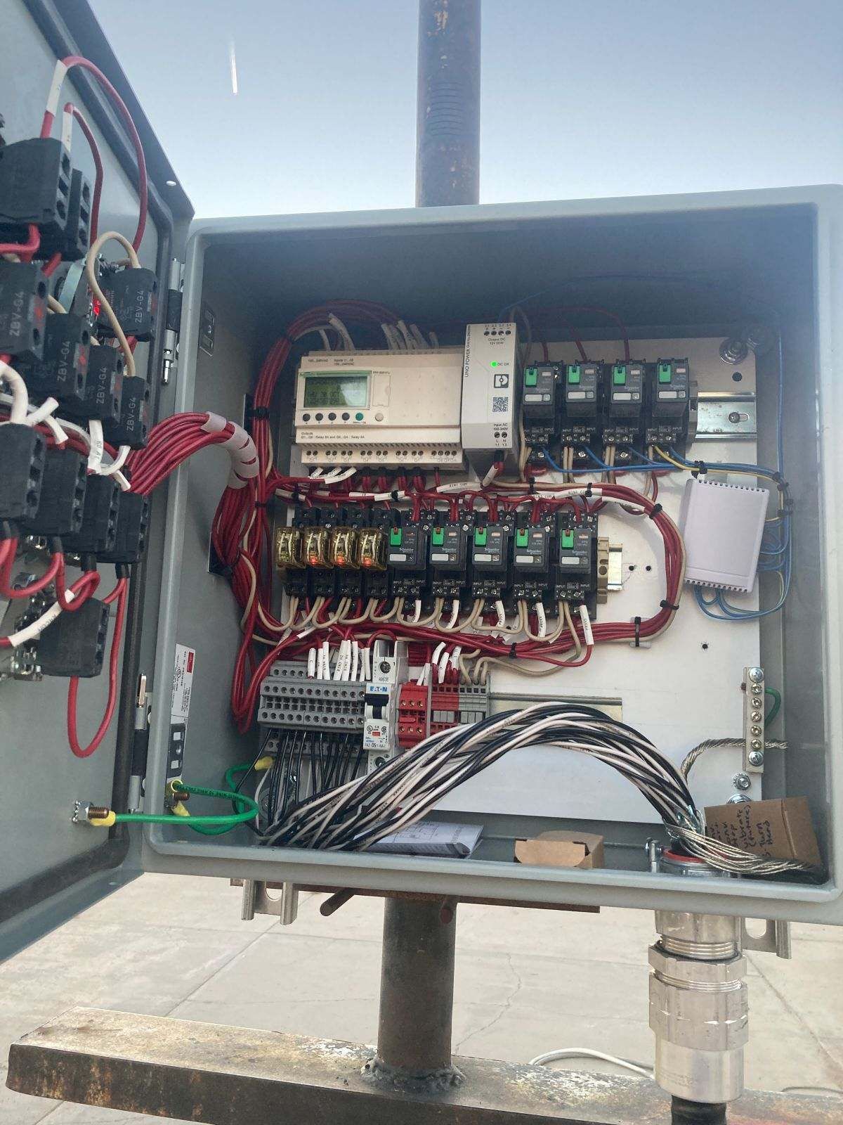 Open electrical control box with wiring, relays, and controller mounted on a pole outdoors.